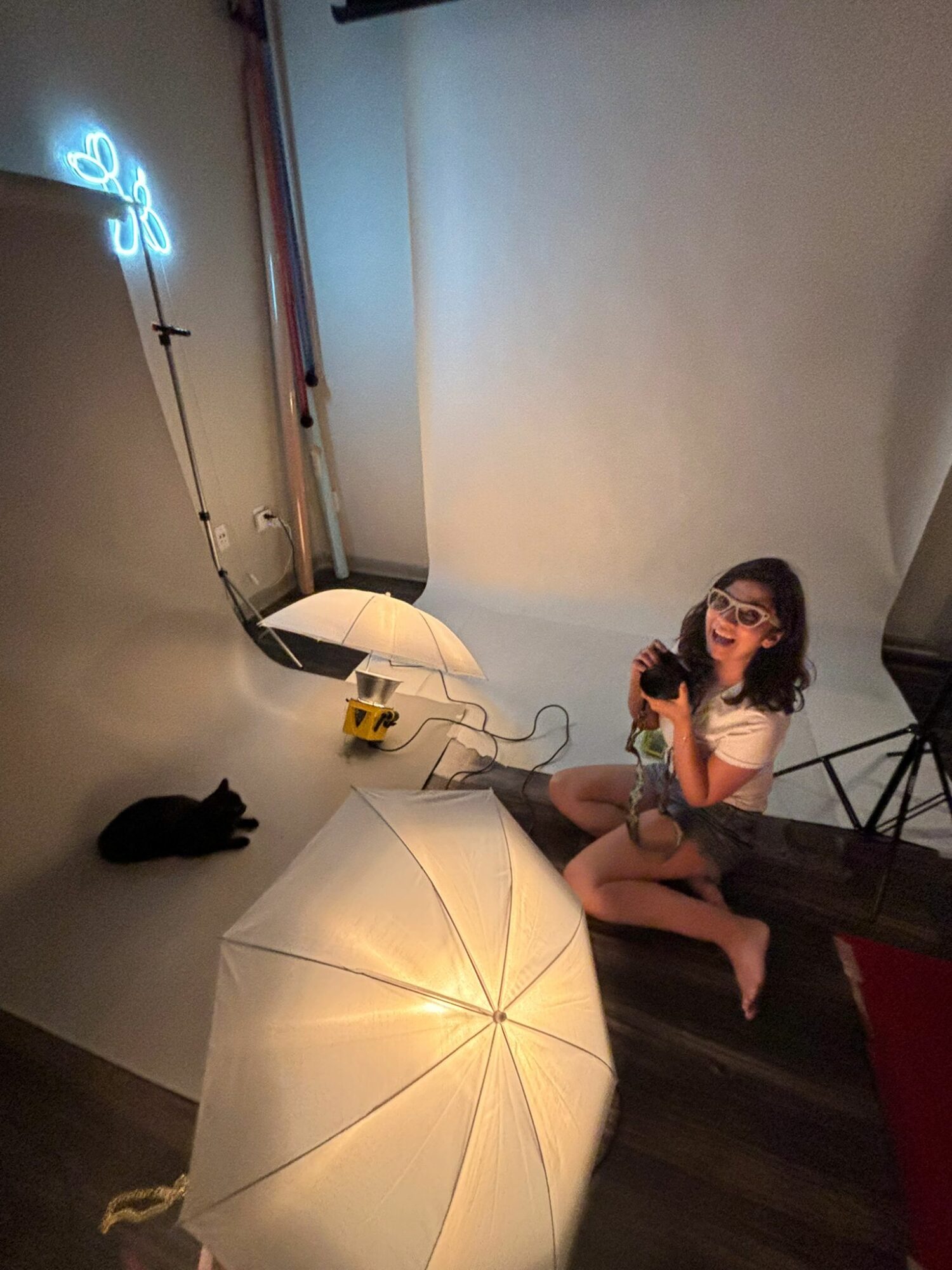 Girl sitting on floor with glasses, holding a phone, surrounded by umbrellas and a neon light, in a room with a white wall.