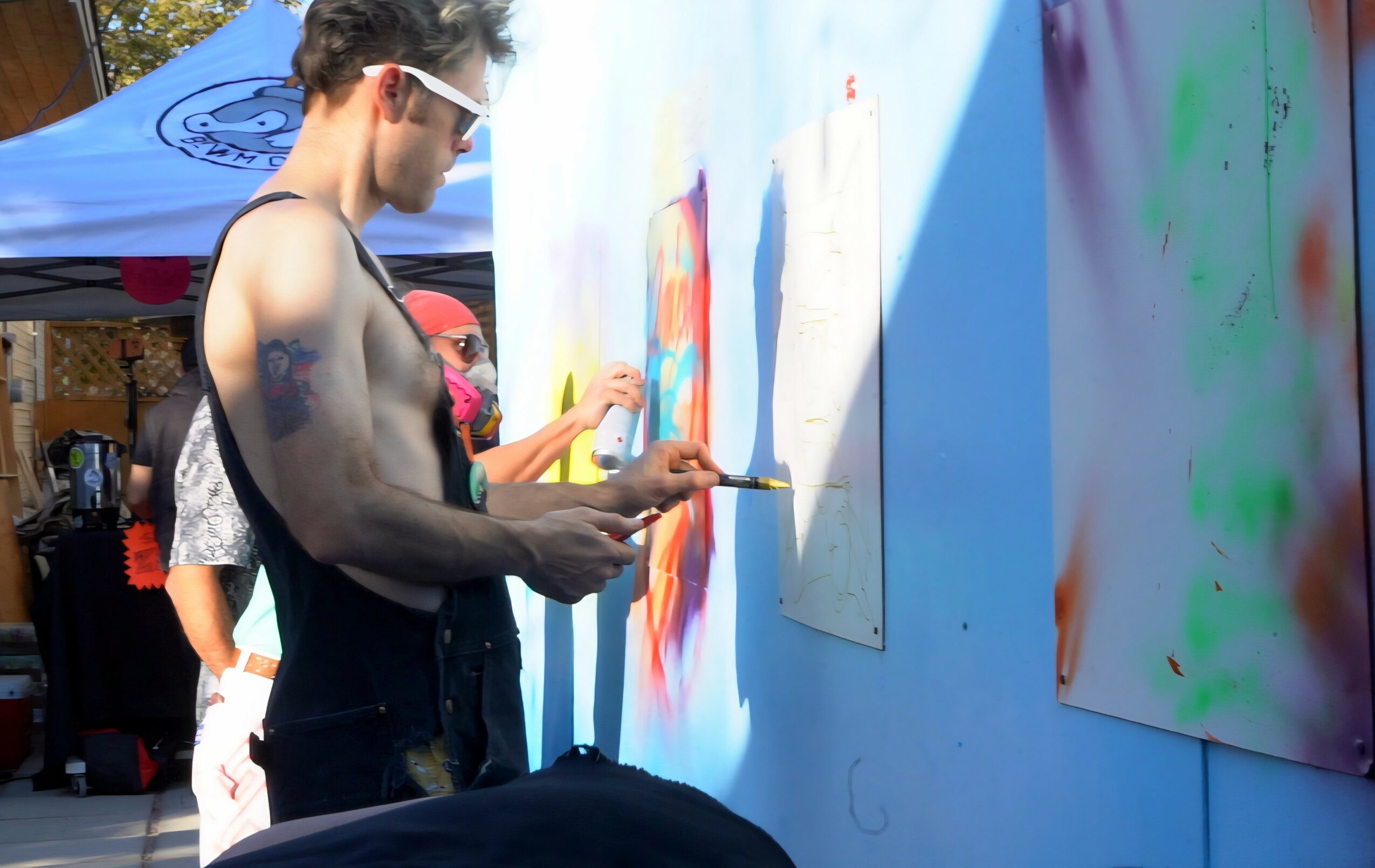Man painting on a large canvas outdoors, with other people and tents visible in the background.
