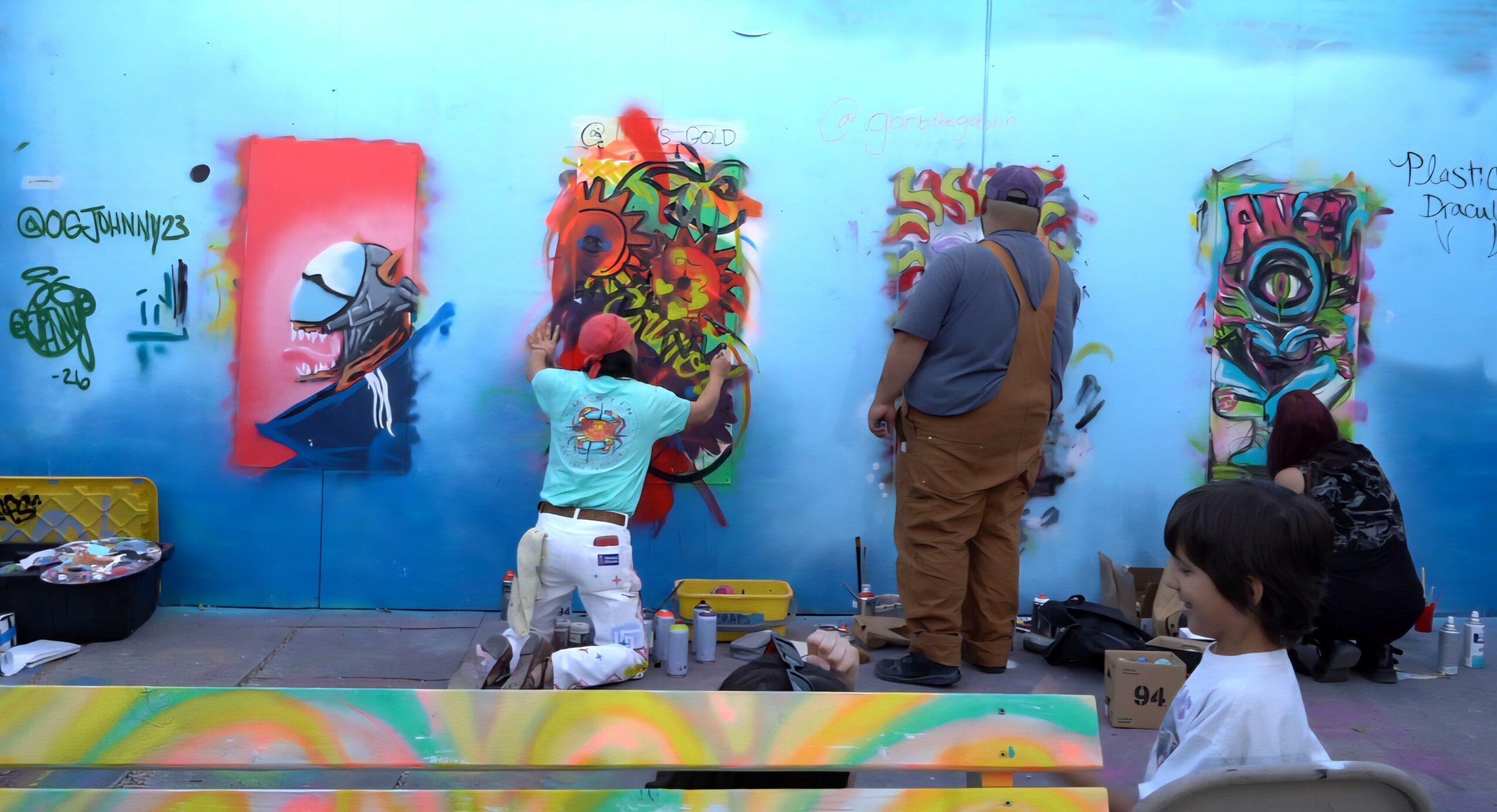 People painting colorful murals on a blue wall, children and adults involved in artwork, with supplies nearby.