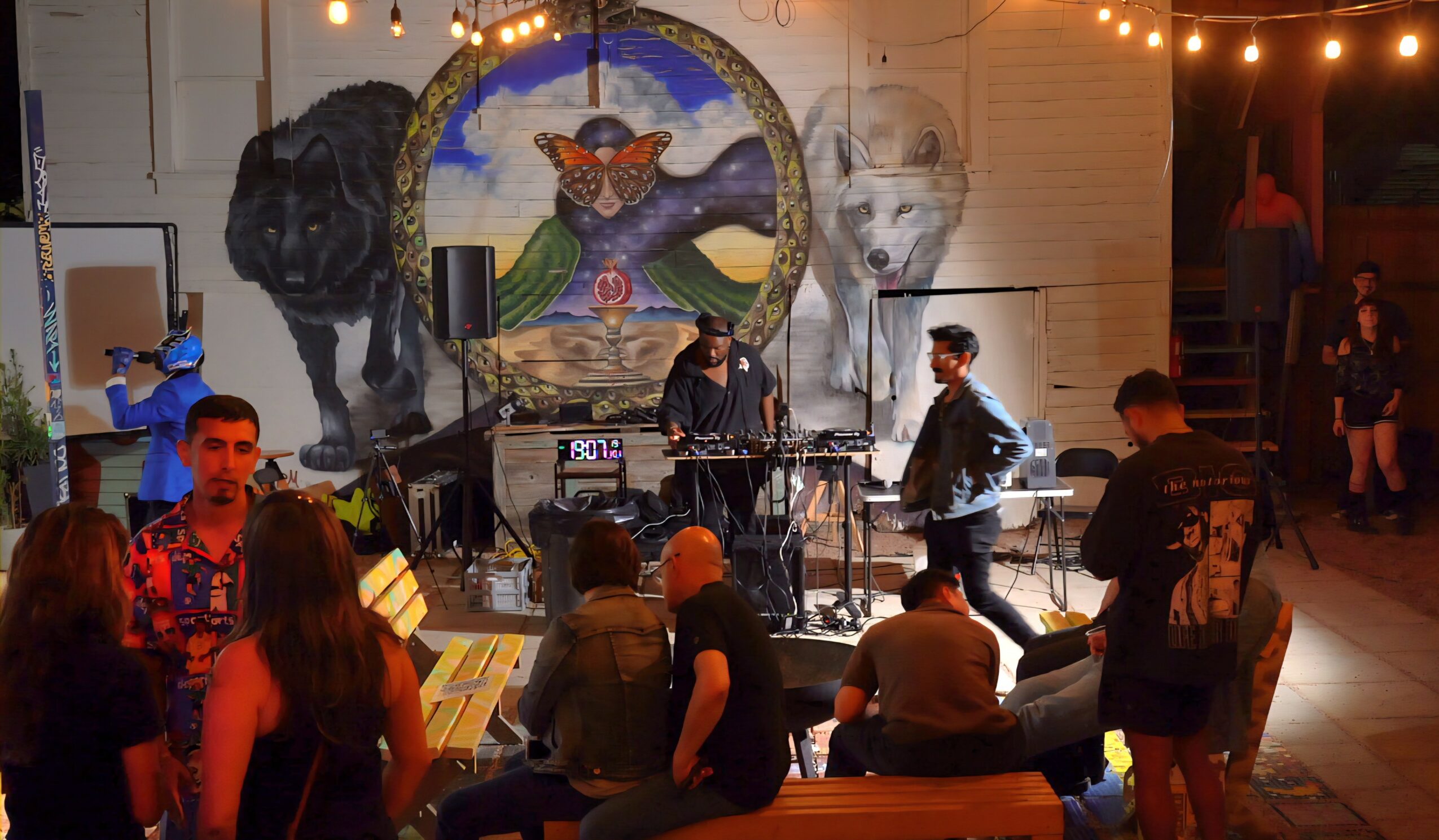People gather around a stage with a mural of a mountain, trees, and a cat, performing music at night.