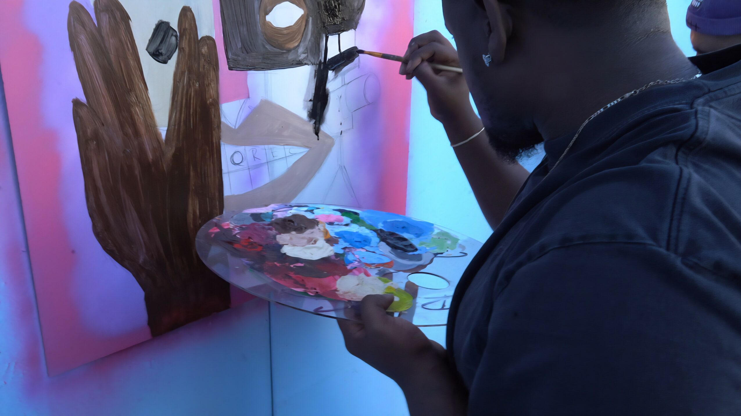 Person painting a mural with a palette, focusing on a hand-shaped tree and abstract shapes, in a colorful setting.