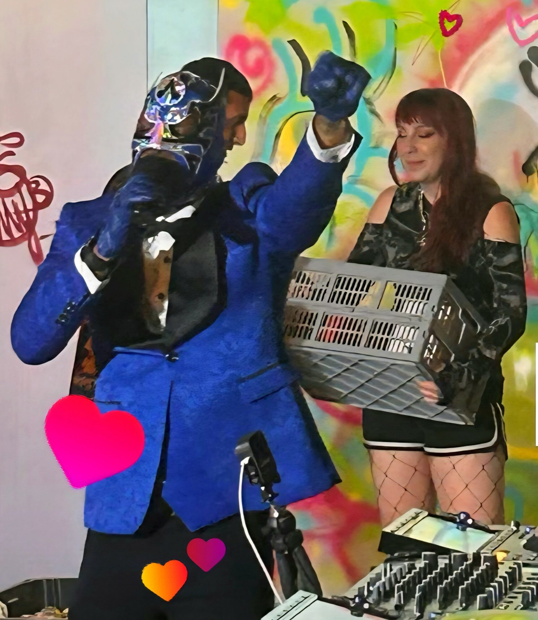 Two people on stage, one in a blue jacket and mask, the other with long hair holding a crate, colorful background.
