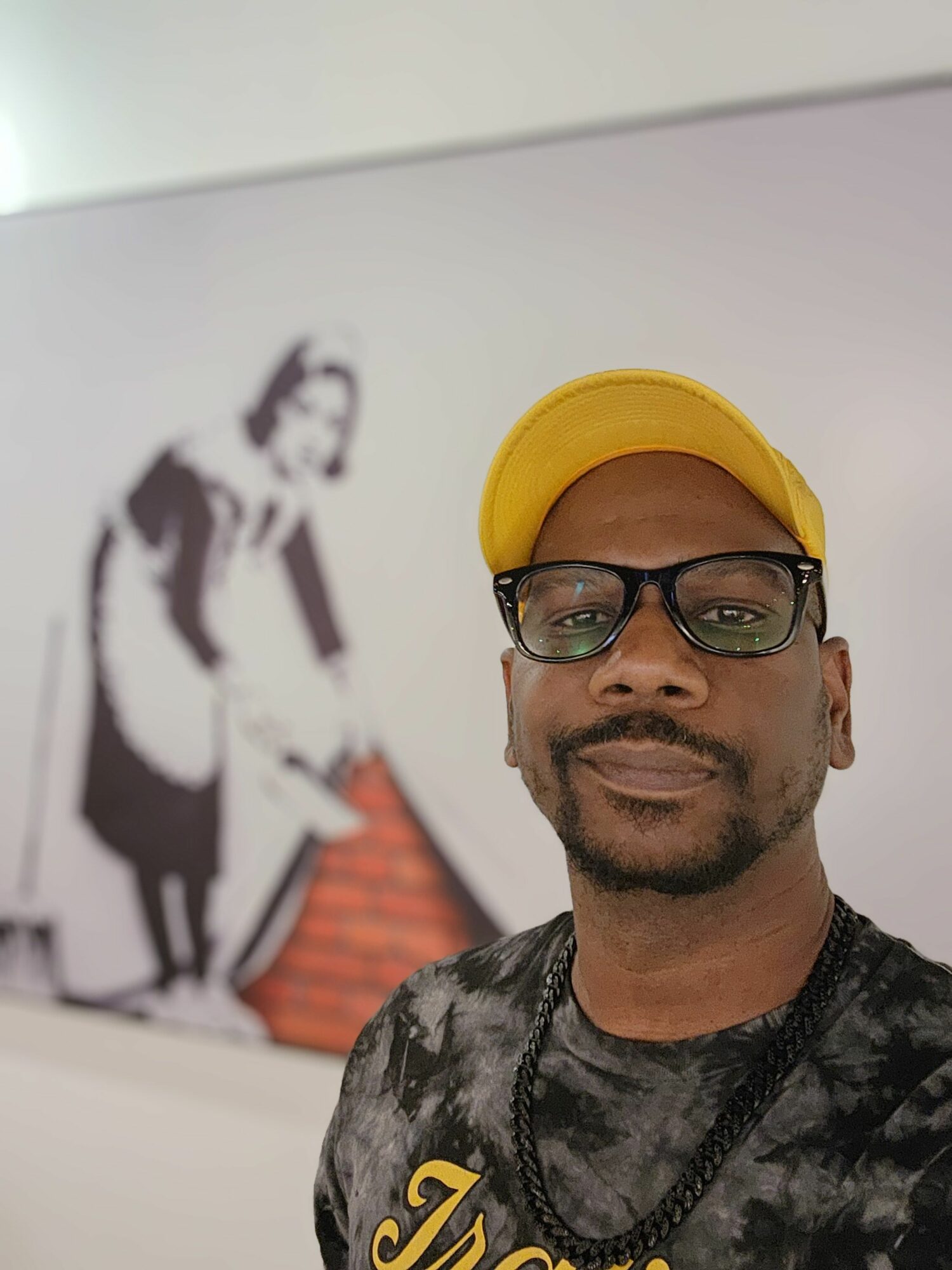 Man wearing glasses and a yellow cap, looking at the camera, with a black and white artwork in the background.