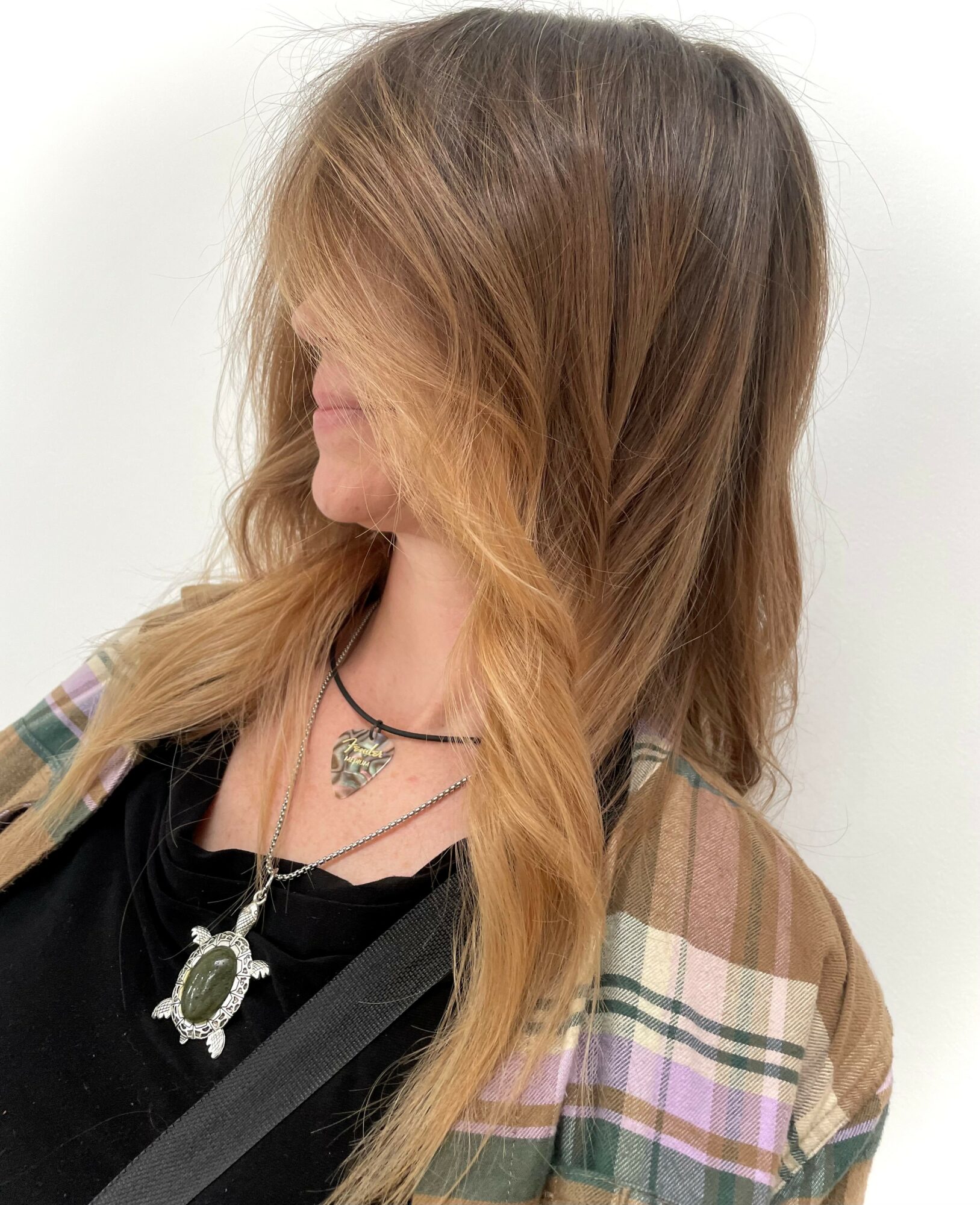Person with long, wavy hair wearing layered necklaces and a plaid jacket, facing left against a plain background.