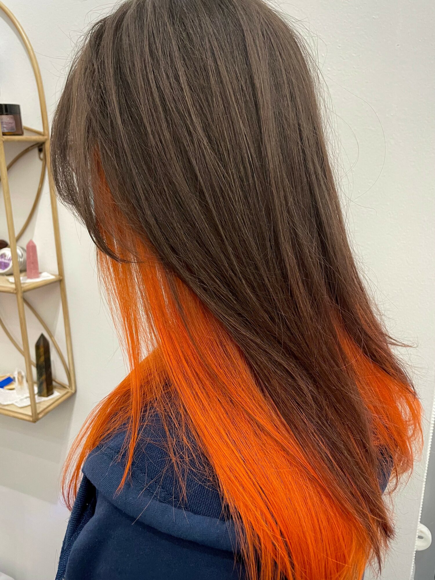 Person with long brown hair and orange tips, wearing a dark blue top, standing indoors near a shelf.
