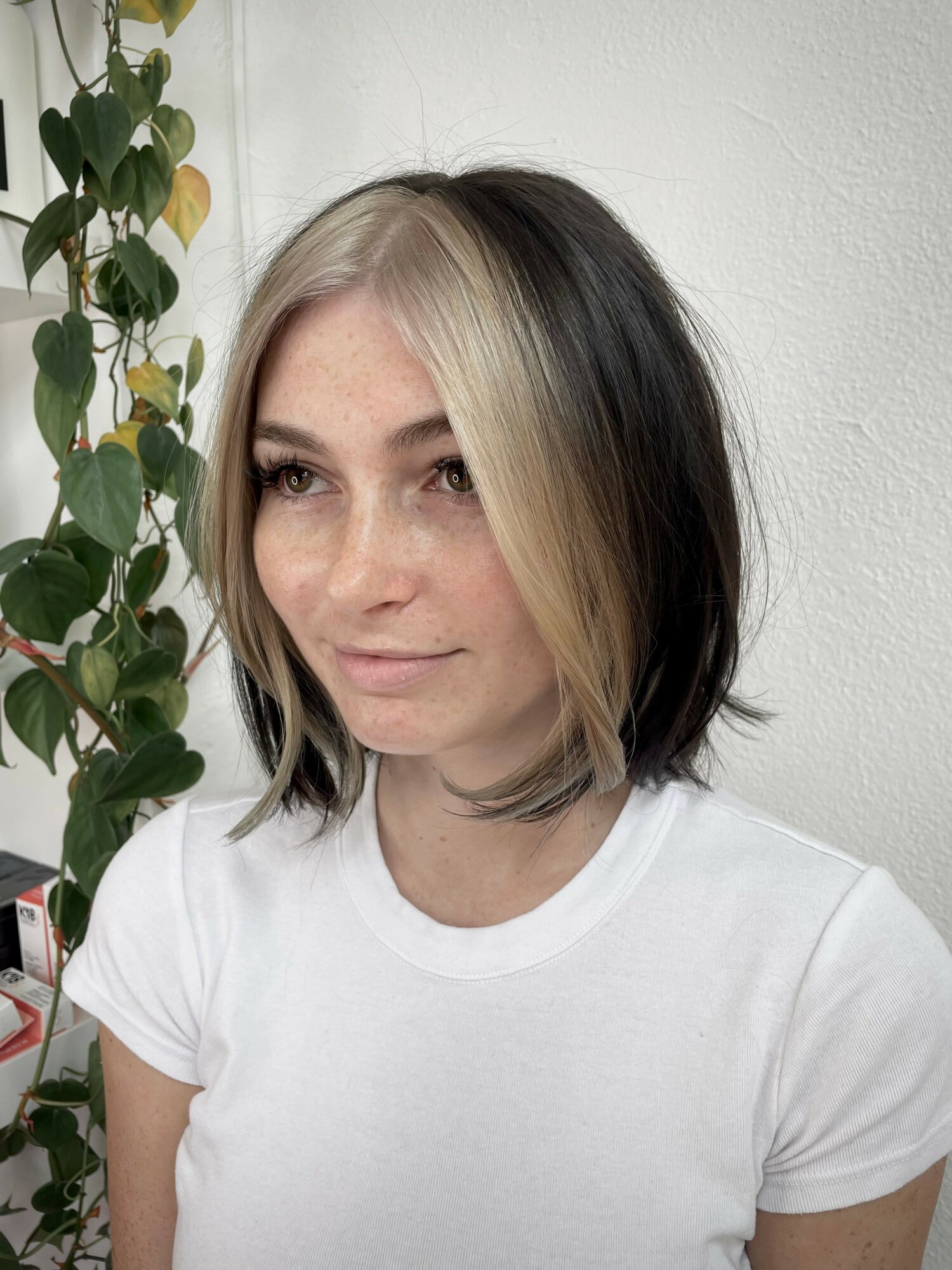 Woman with shoulder-length hair, blonde on one side, black on the other, wearing a white t-shirt, standing near a plant.