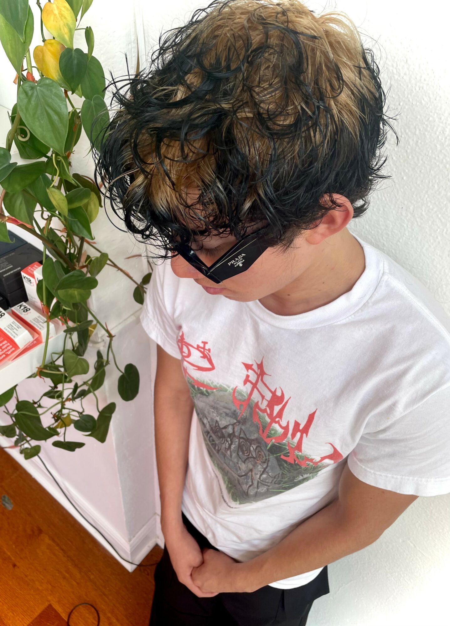 Young person with curly hair wearing sunglasses and a white T-shirt, standing next to a plant and a white wall.
