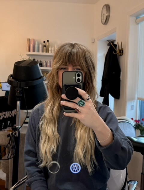 Person with long wavy hair taking a mirror selfie in a room with shelves and a clock, holding a phone with a pop socket.