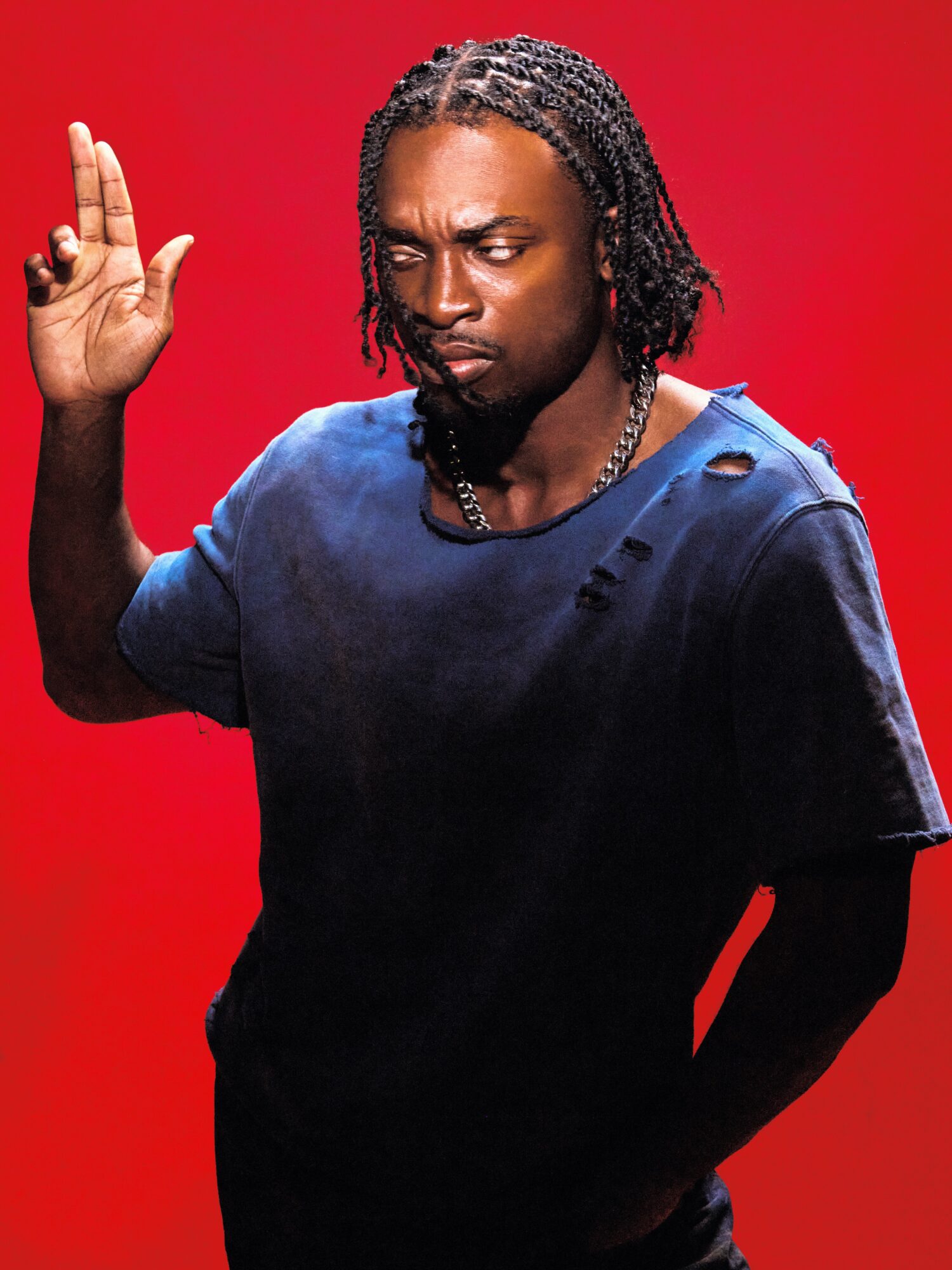 Person with braided hair and a serious expression, raising one hand, against a red background.
