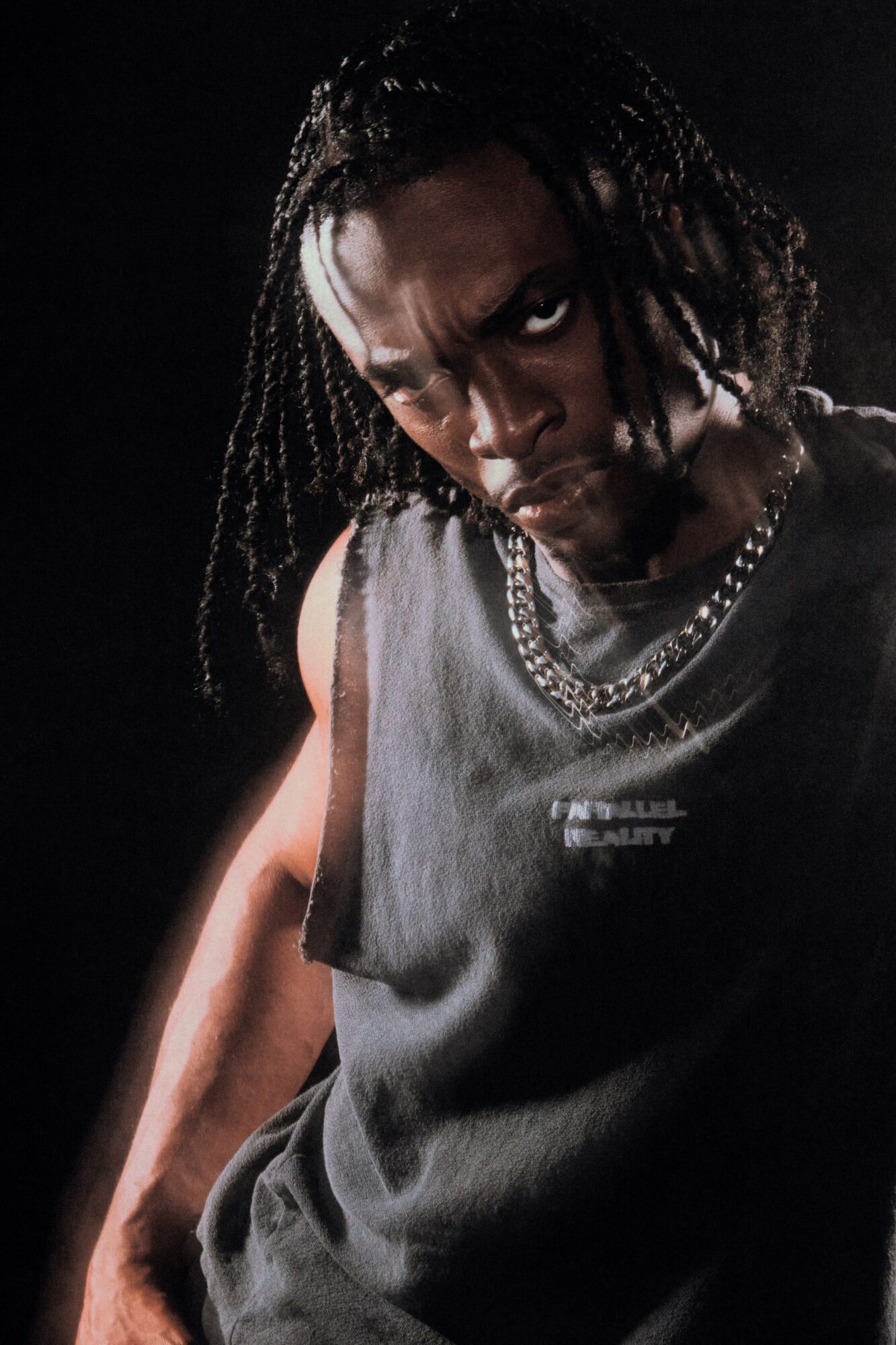 Person with braided hair wearing a sleeveless shirt and layered necklaces, looking intensely at the camera.