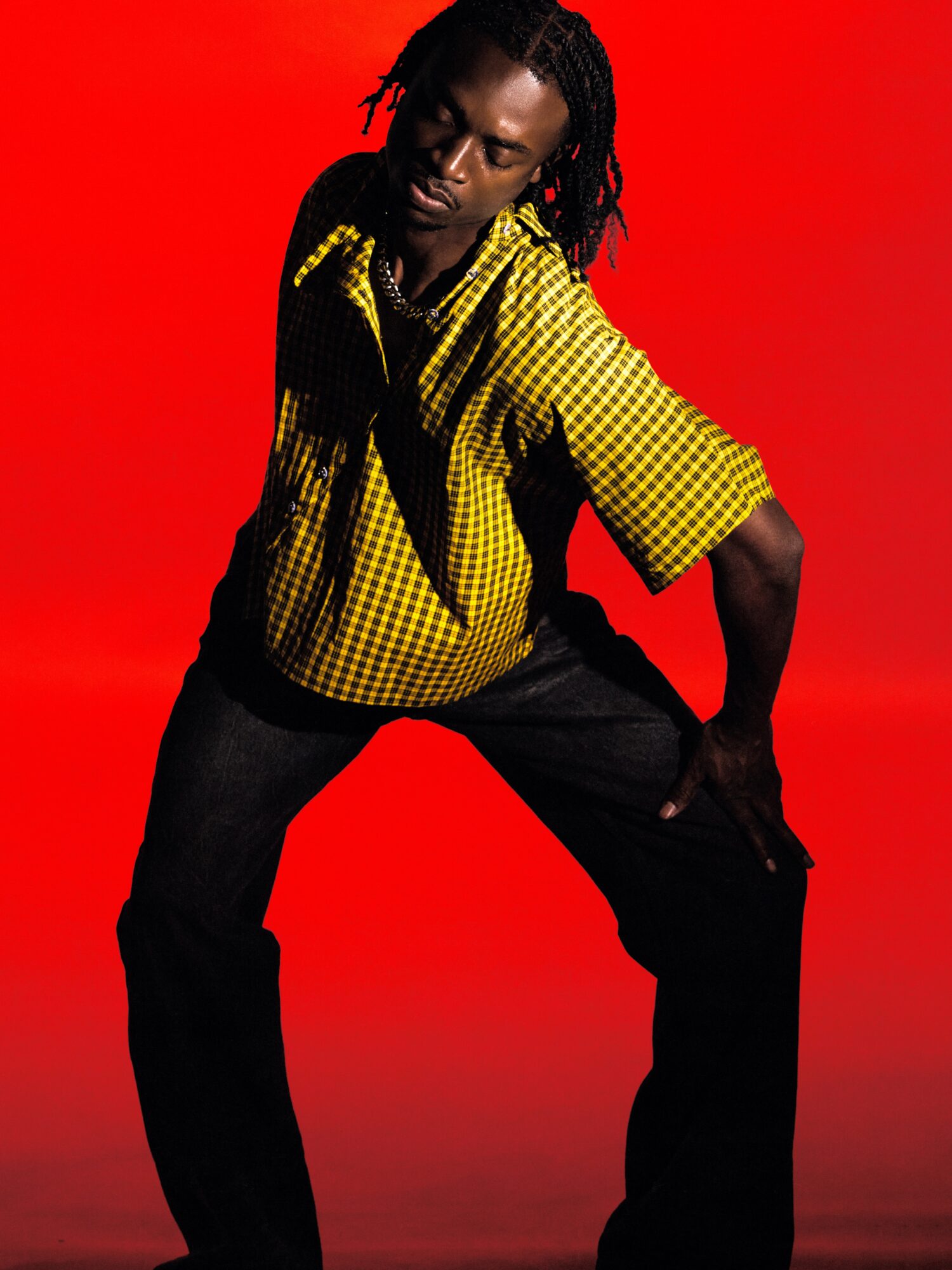 Person with dreadlocks wearing a yellow checkered shirt and black pants, posed against a red background.