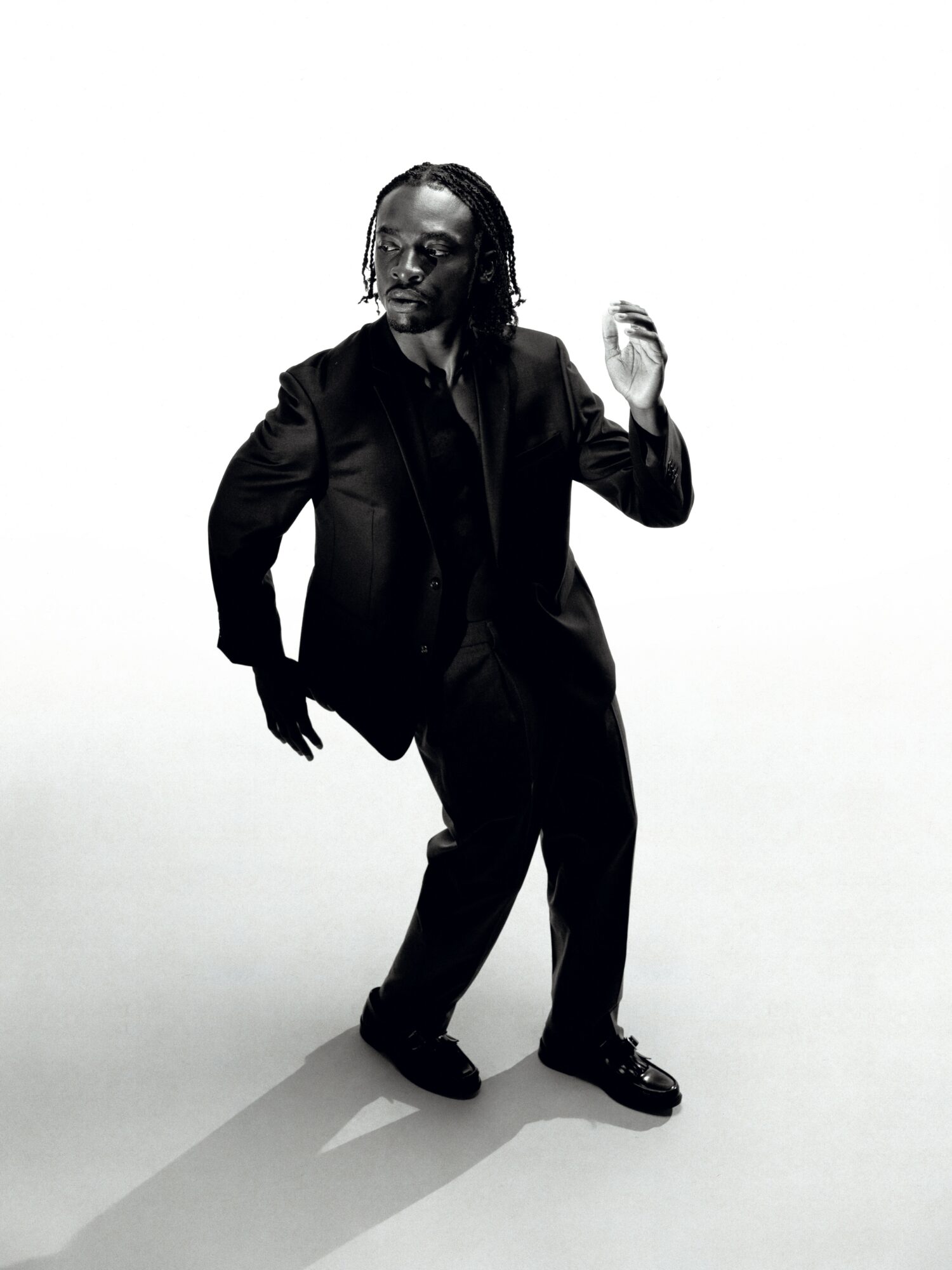 Person with dreadlocks in a suit, standing and gesturing with one hand, against a plain background.