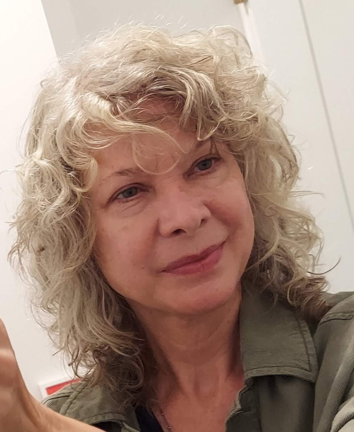 A woman with curly blonde hair smiling, wearing a green jacket, in an indoor setting.