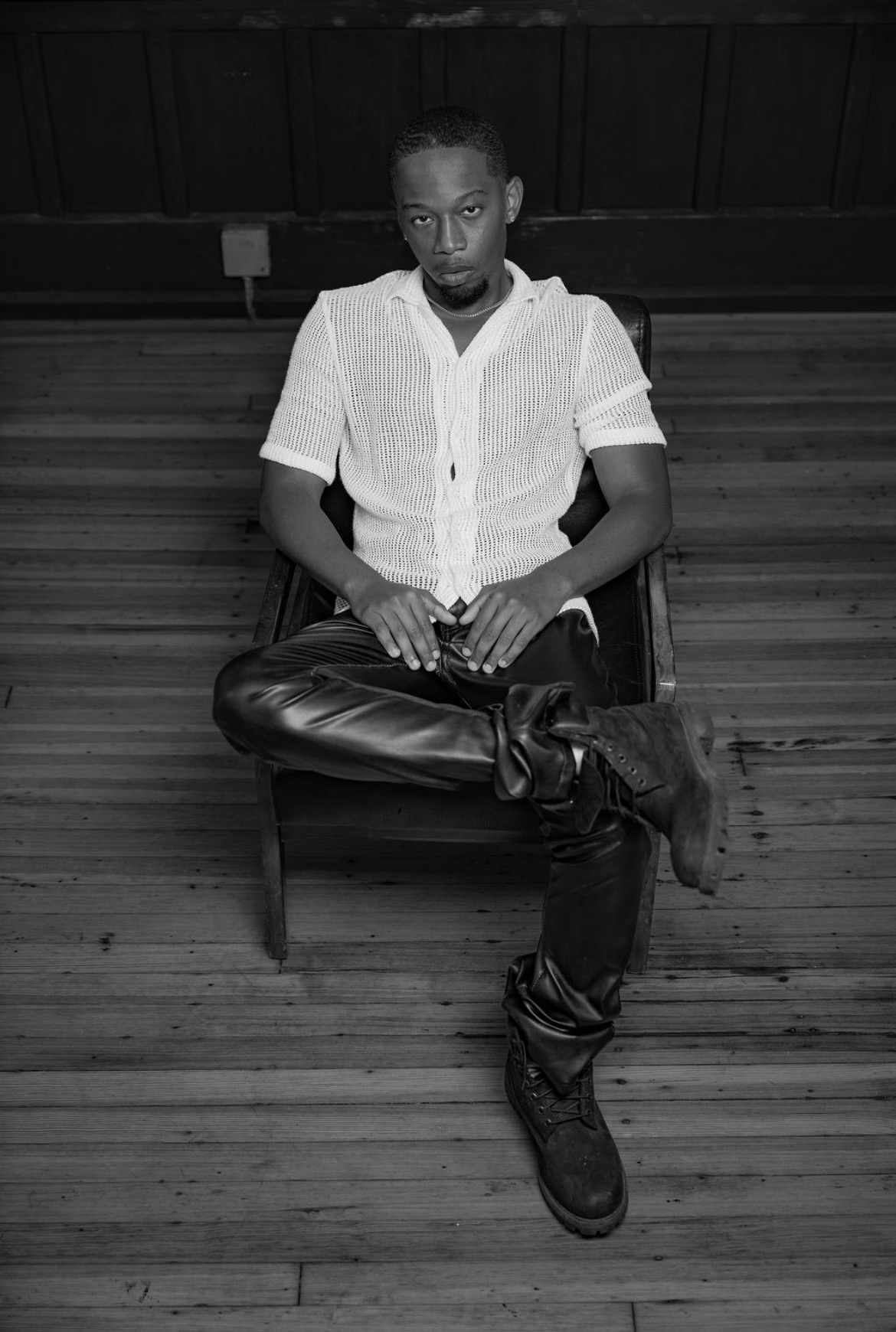 Person sitting on a chair with crossed legs, wearing a white shirt and dark pants, in a room with wooden floor and ceiling.