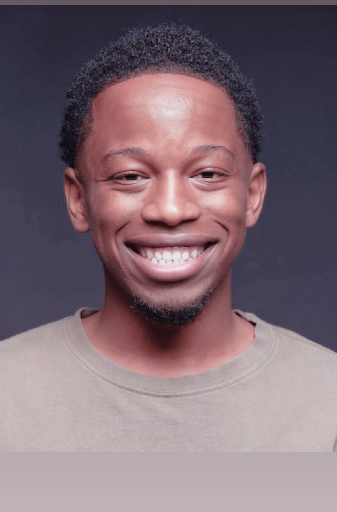 Smiling person with short curly hair and a beige shirt, facing forward, dark background.