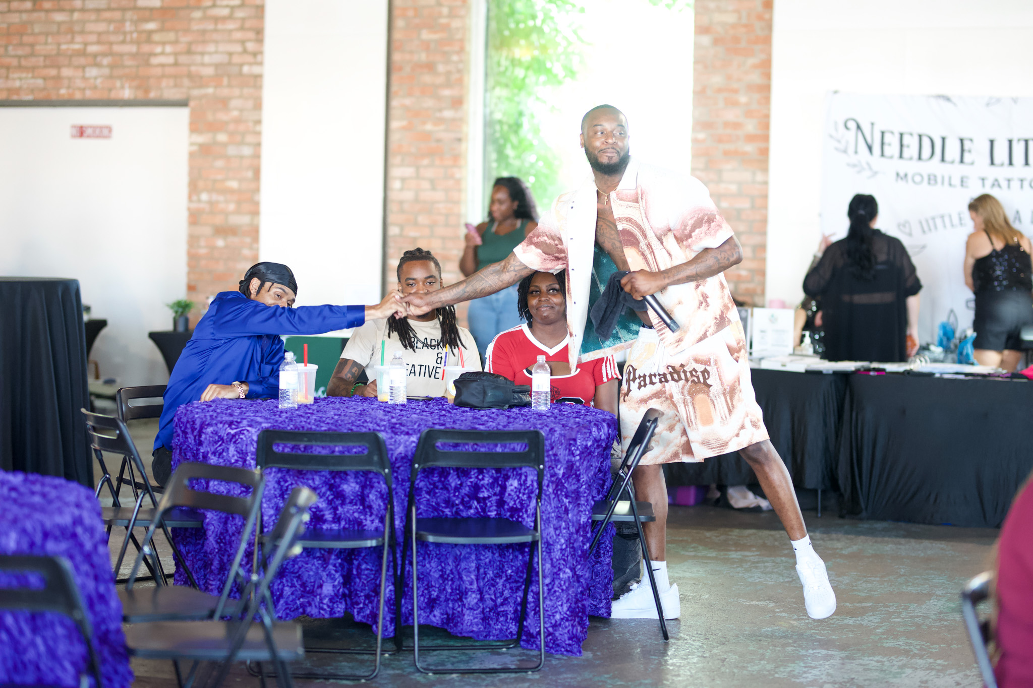 Four people sitting at a table with purple tablecloths, one person standing and holding a tattoo machine, in a large indoor space.