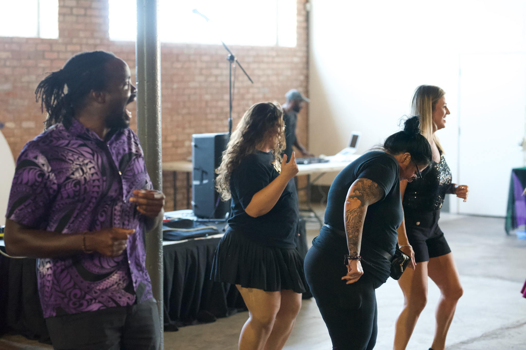 Four women dancing and socializing in a bright indoor space with brick walls and large windows.