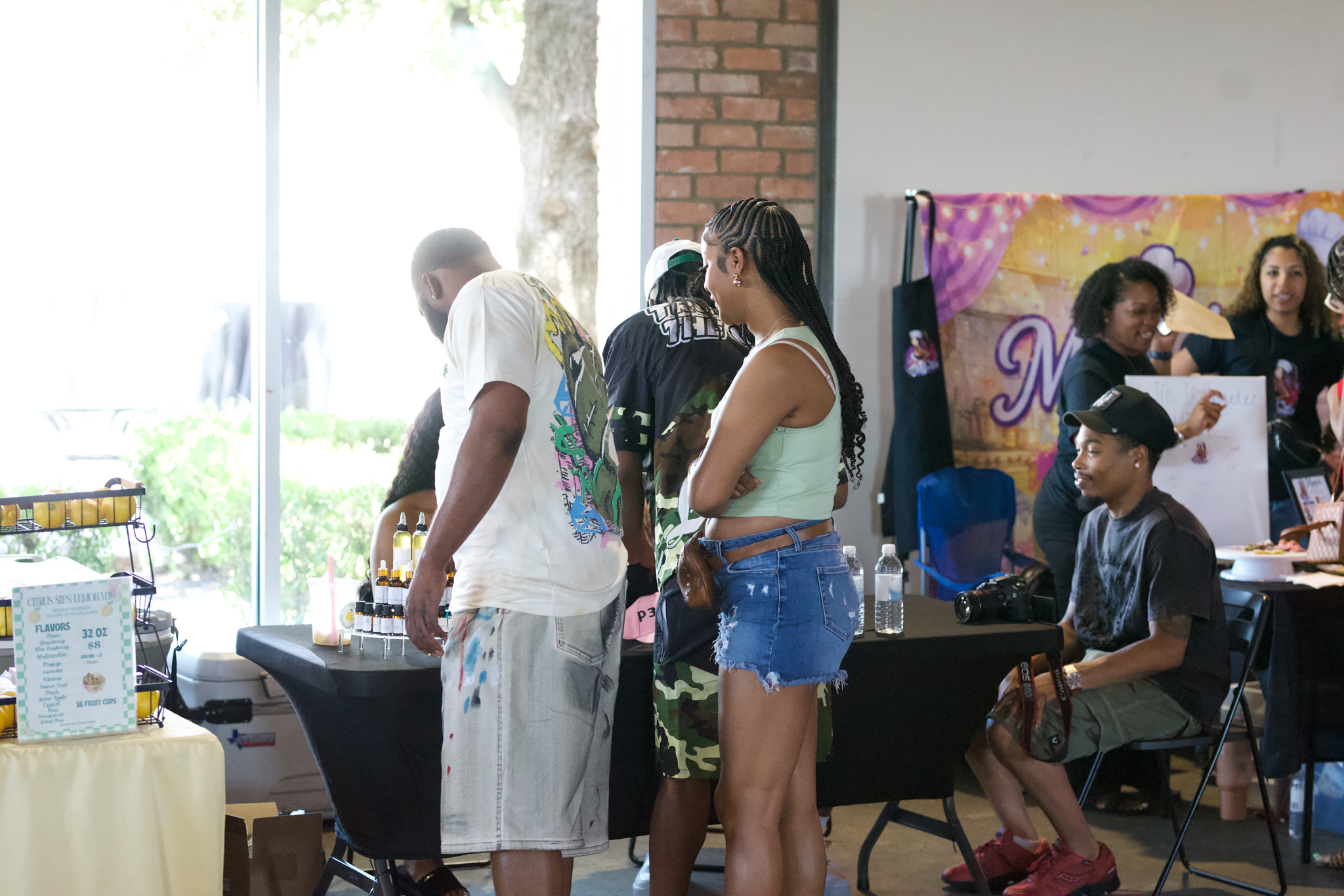 Group of people gathered indoors near a table with a window in the background, some standing and some sitting.