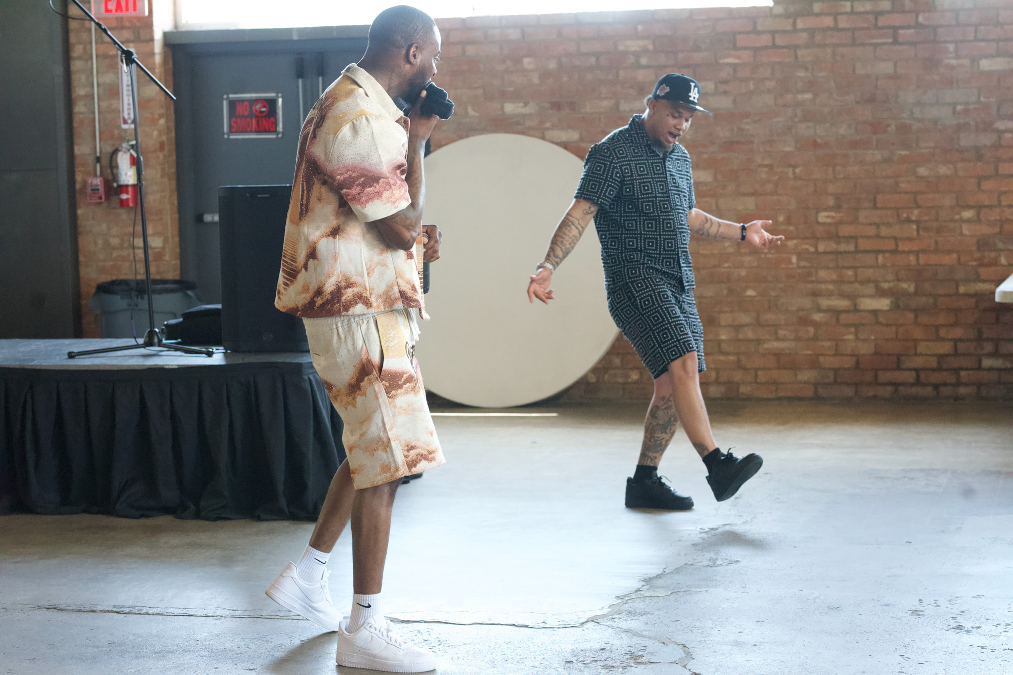Two people dancing indoors with a brick wall background, one wearing a patterned outfit and the other in a dark shirt and shorts.