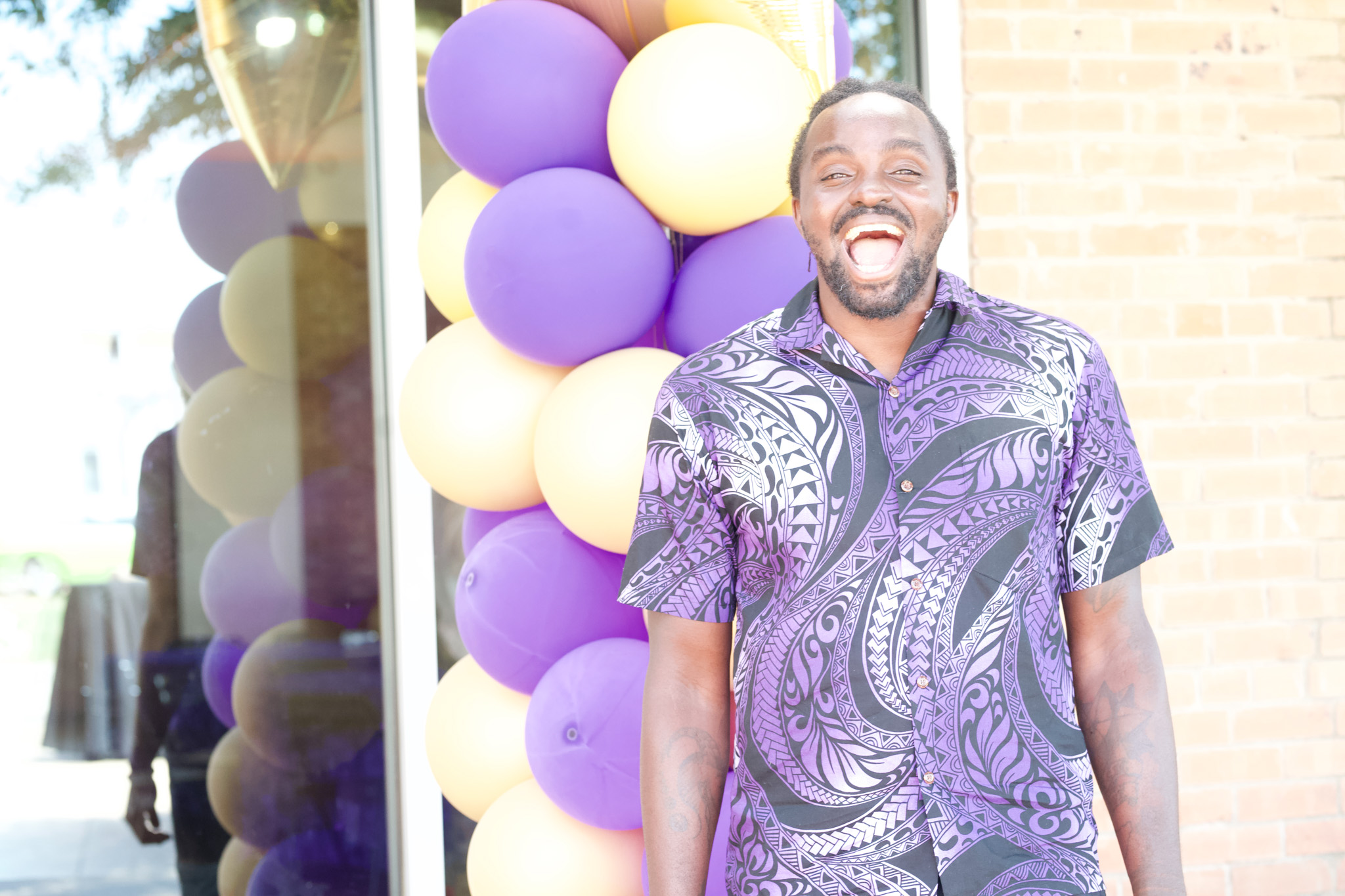 Man smiling outdoors near colorful balloons, wearing a patterned purple shirt, with a brick wall in the background.