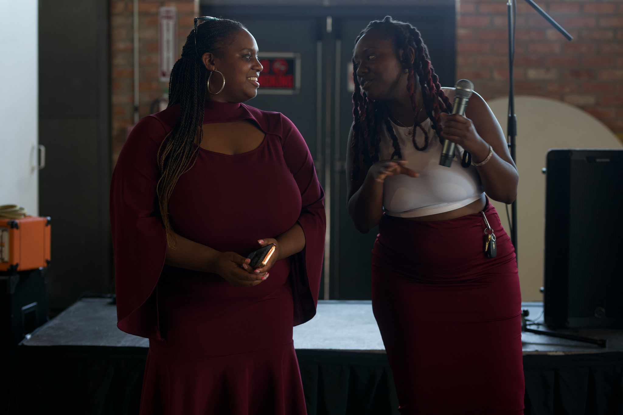 Two women with braided hair and dark skin talking on a stage with a brick wall background.