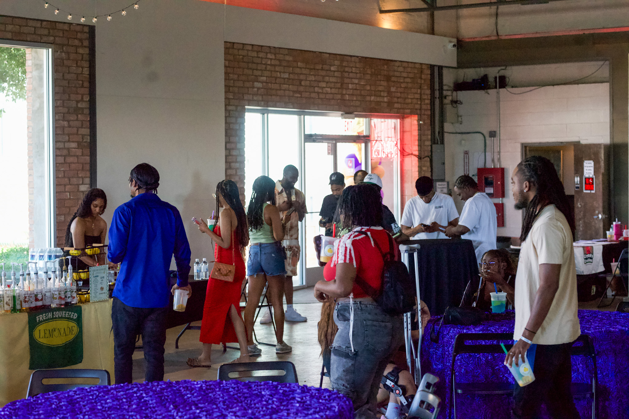People gather inside a room with brick walls, some standing near tables, others walking or talking, with a window and door in background.