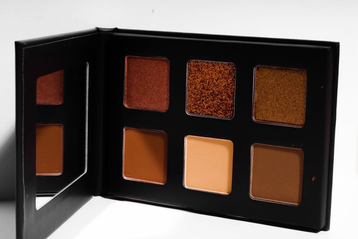 Open eyeshadow palette with six shades in black case, including browns, oranges, and golds, arranged in two rows.
