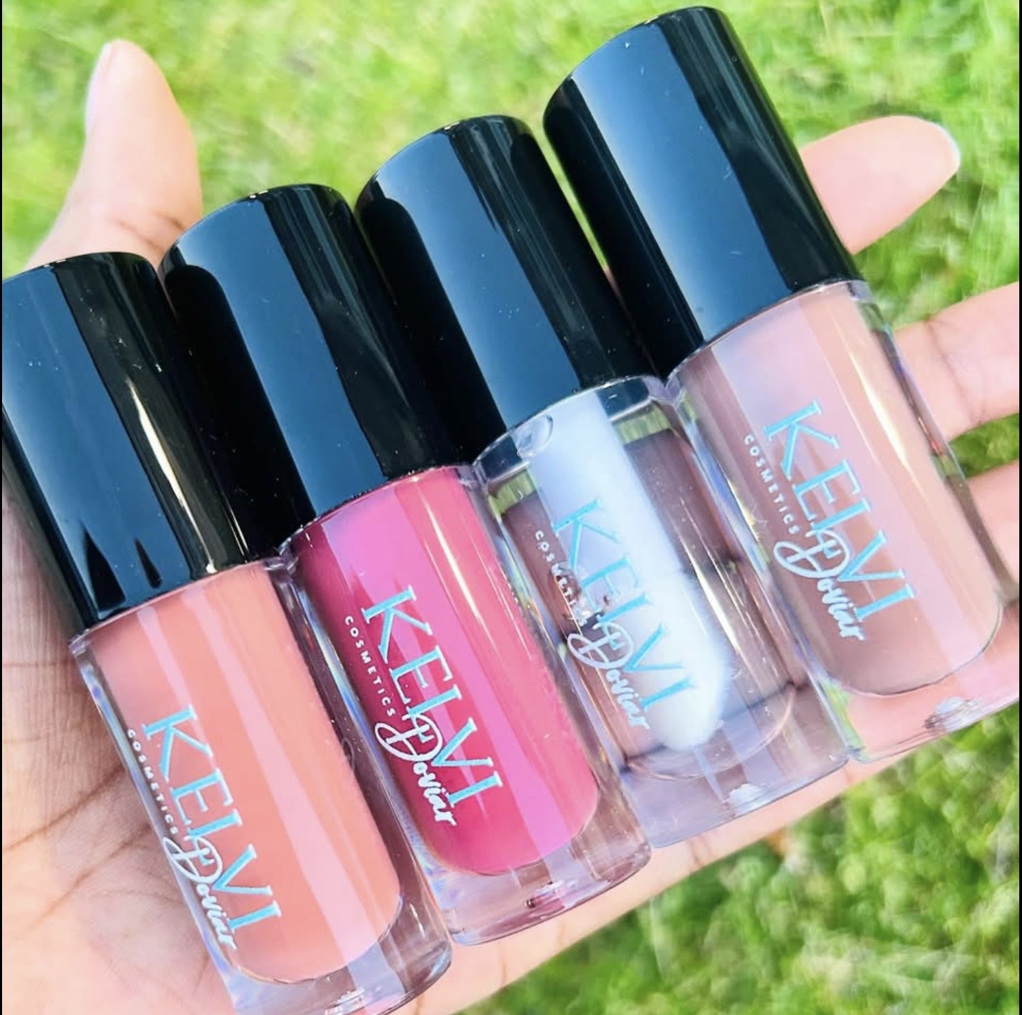 Hand holding five bottles of nail polish with black caps, pink and blue shades, outdoors on grass