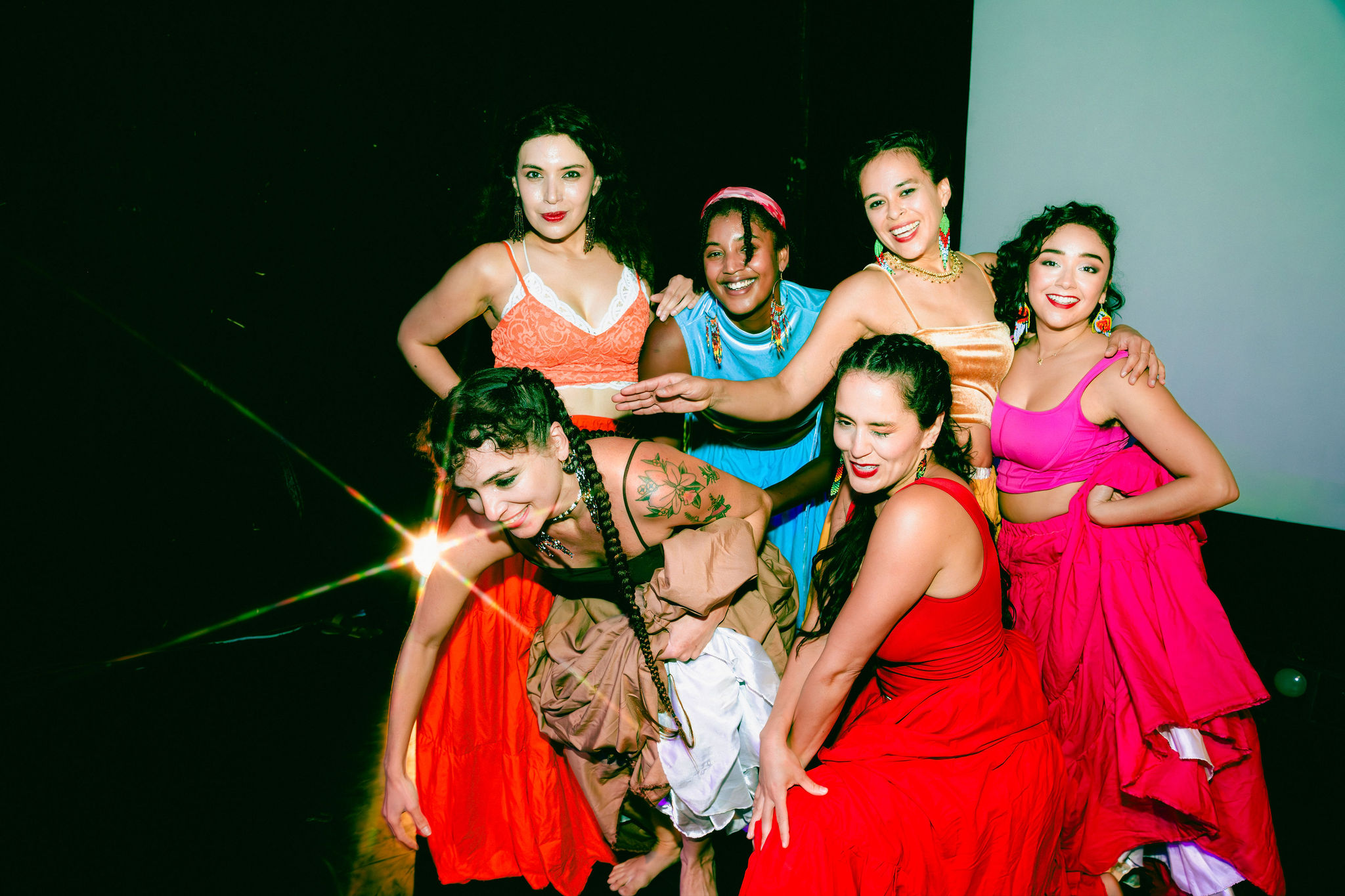 Group of six women in colorful costumes smiling together, some with arms around each other, against a dark background.