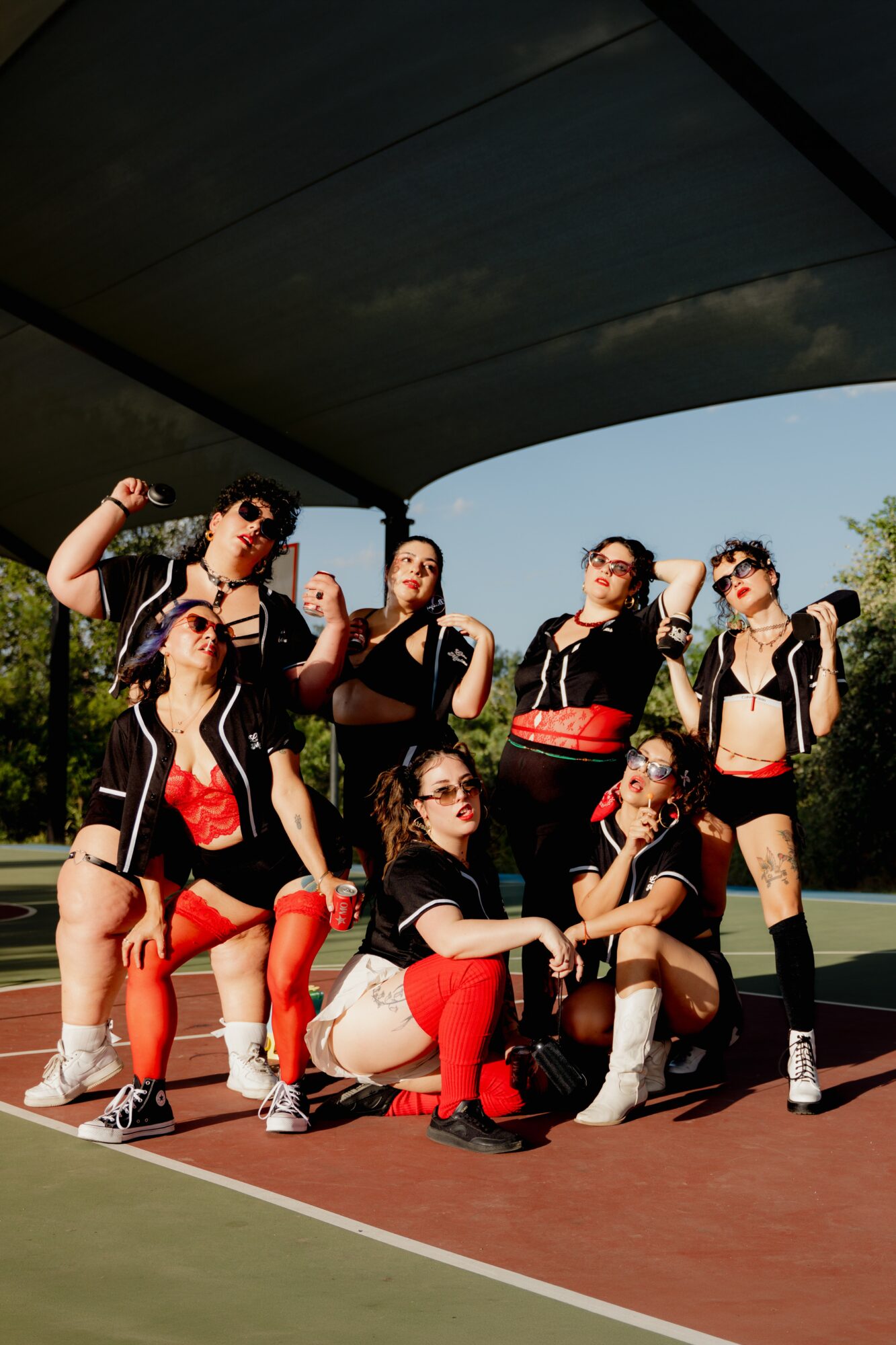 Group of seven women posing on a tennis court under a canopy, wearing sporty outfits with sunglasses, some holding tennis rackets.