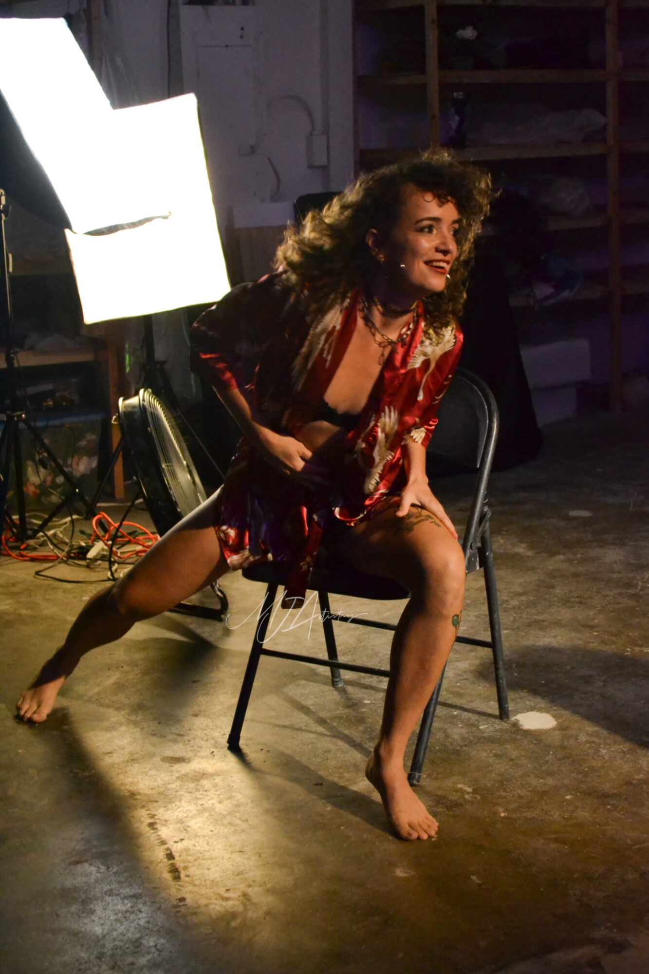 Performer with curly hair in a red and black costume, sitting on a chair with legs apart, on stage.