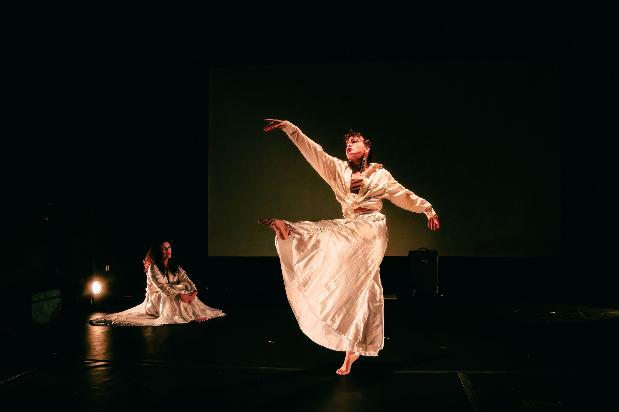 Two dancers perform on stage, one in mid-air with arms extended, the other sitting on the floor, both in flowing costumes.
