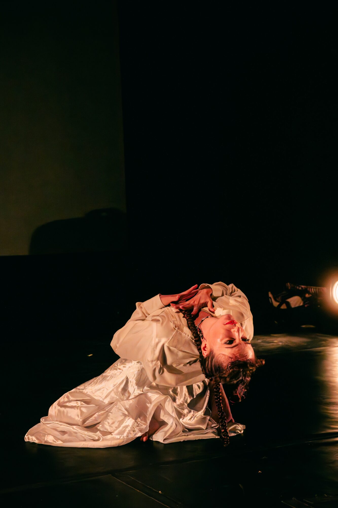 Person lying on the floor with head resting on arm, wearing a white garment, in a dark room with a light source nearby.
