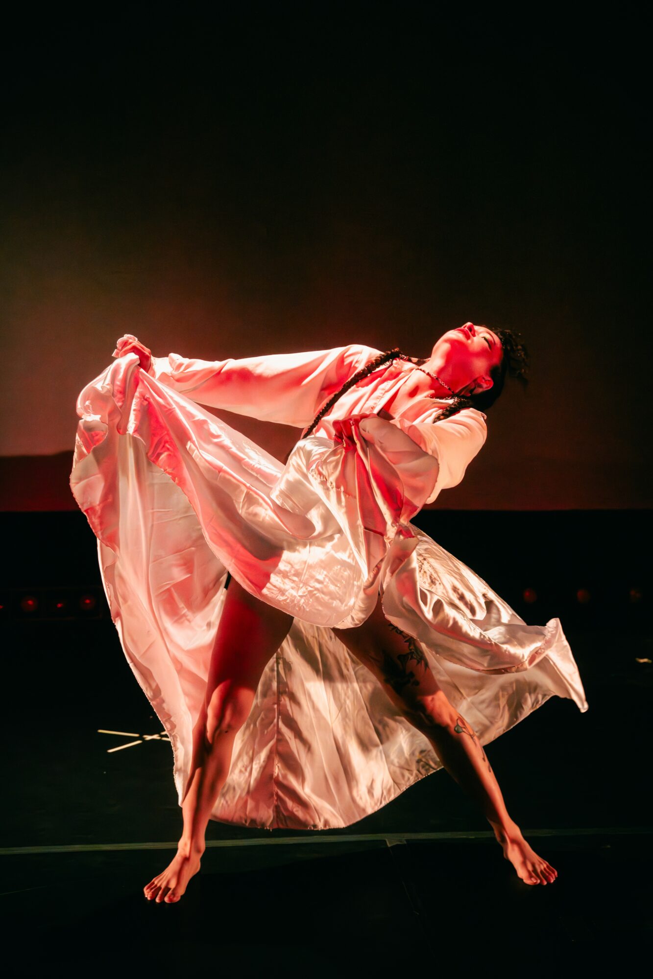 Two dancers perform on stage, one lifting the other, both wearing flowing, shiny costumes, with a dark background.