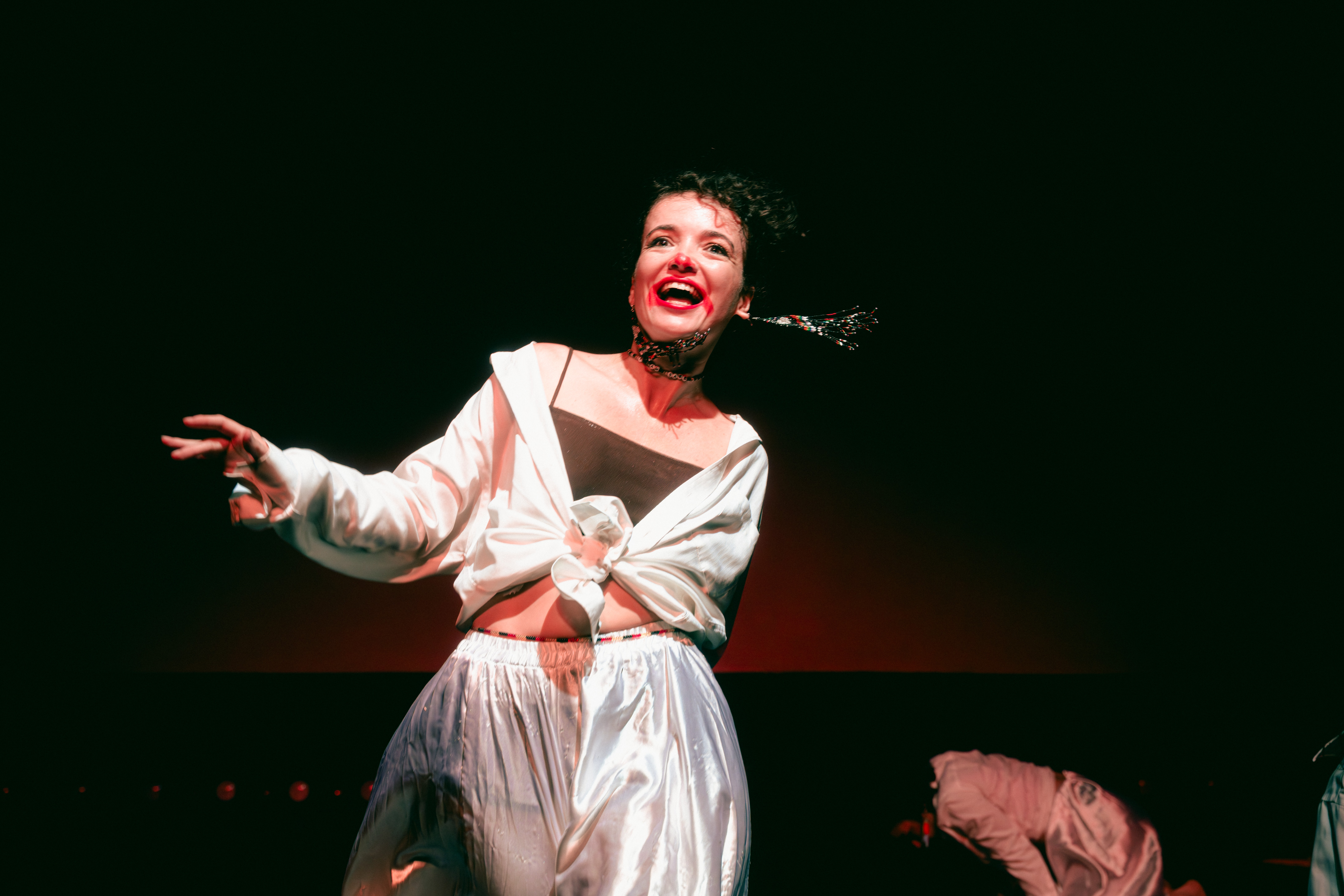 A woman smiling and gesturing with her hand, wearing a white outfit, against a dark background.