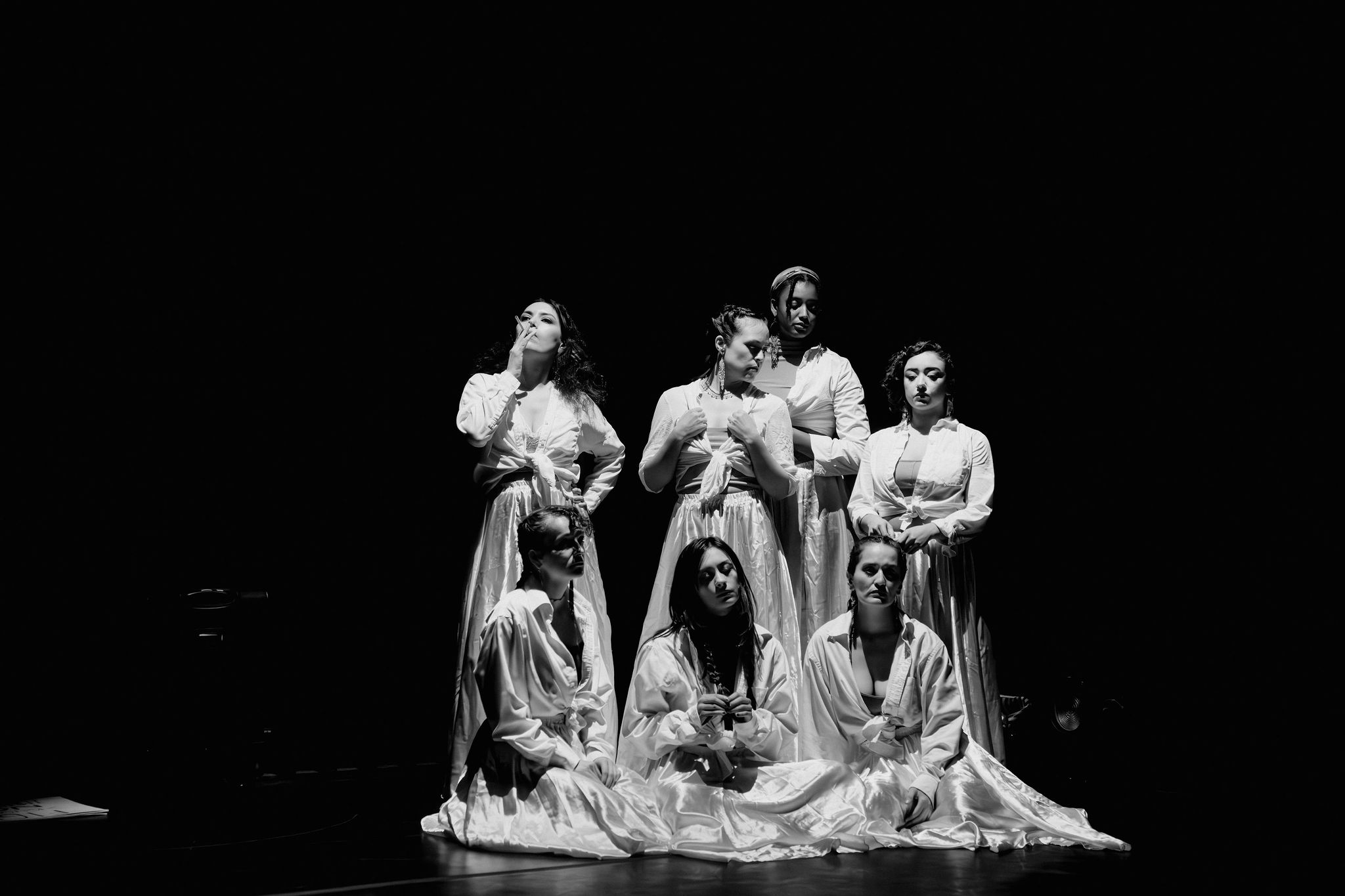 Seven people in costumes pose on stage with a dark background, some standing and some sitting, in a theatrical scene.