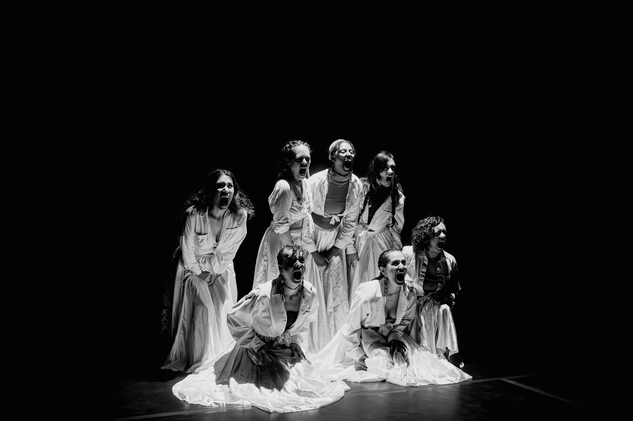 Group of seven women in dresses performing on stage with dark background, some kneeling and some standing, expressive poses.