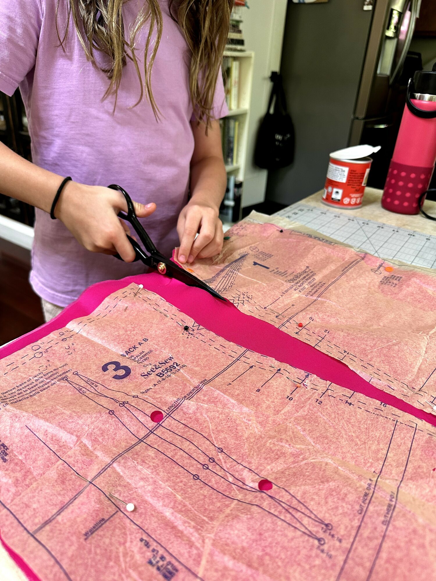 Person cutting pink fabric on a table with sewing pattern, scissors in hand, in a room with shelves and supplies.