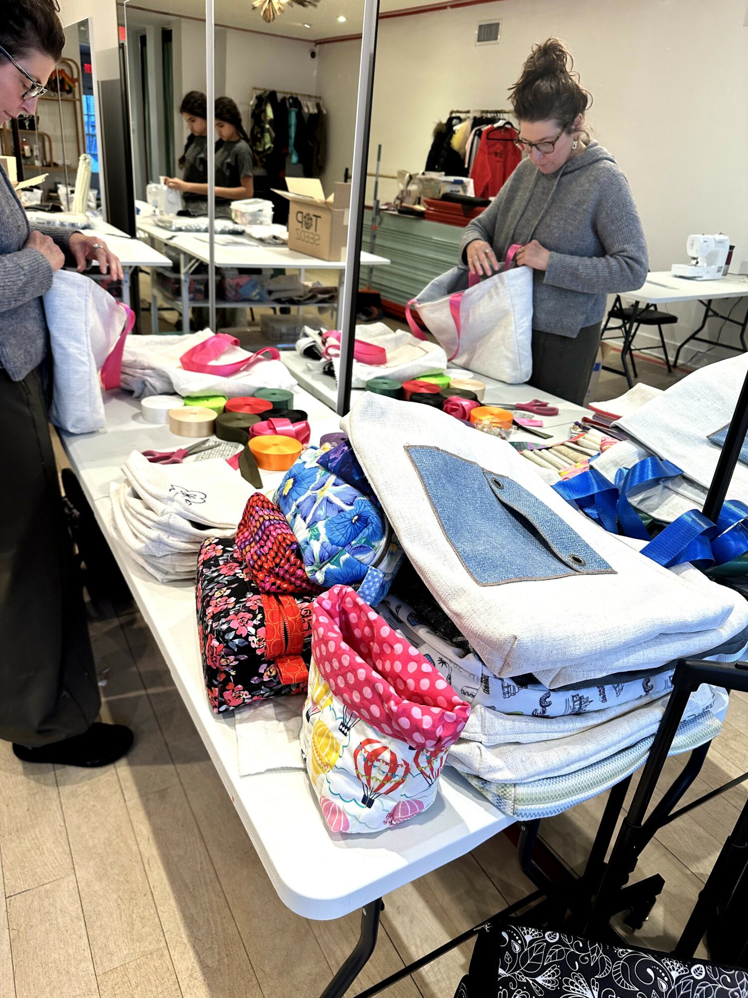 Person in glasses and gray hoodie working at a table with sewing supplies and fabric, in a room with mirrors and clothing racks.