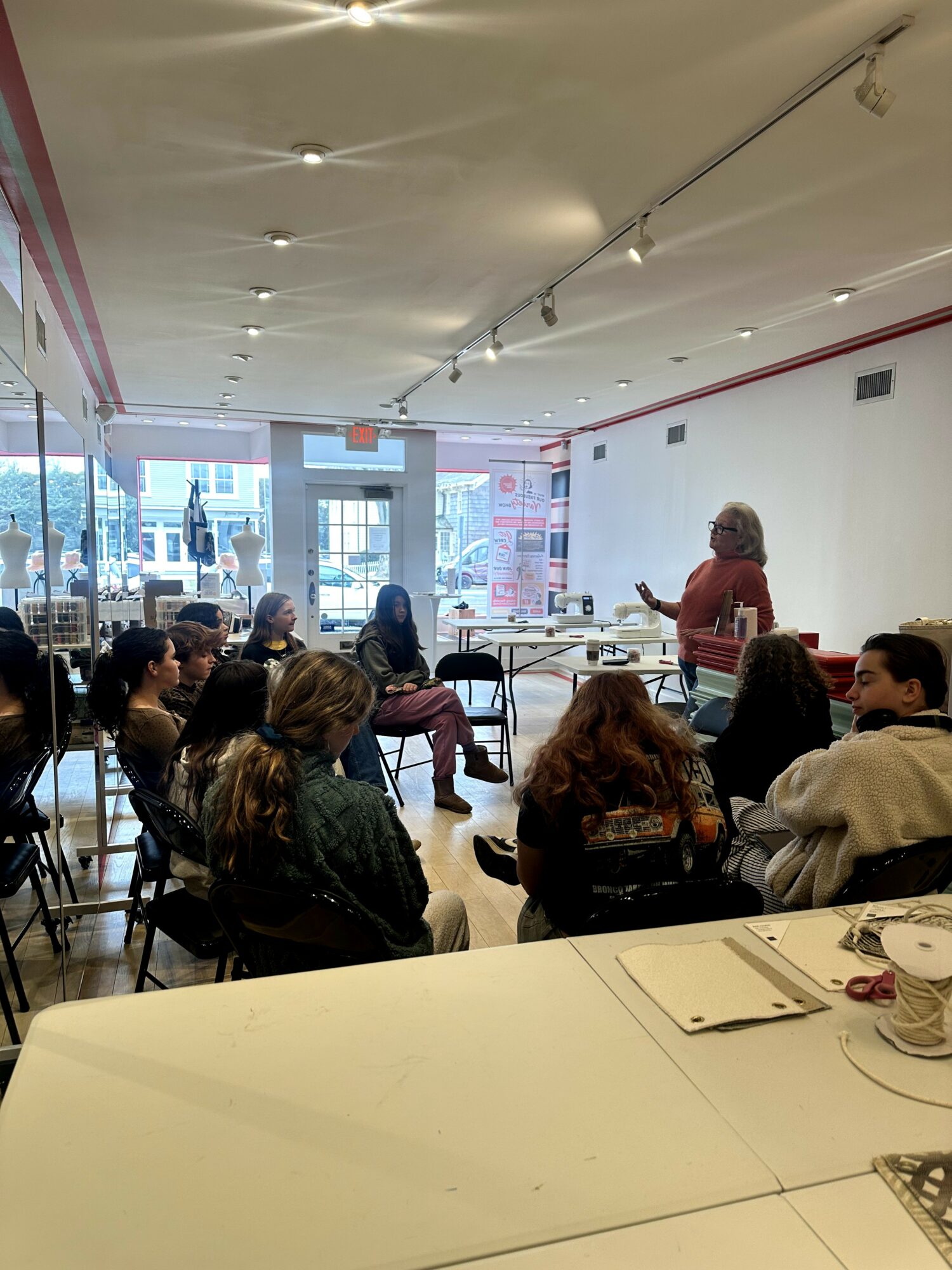 Group of people seated in a room listening to a woman speaking at the front, with large windows and a white wall.