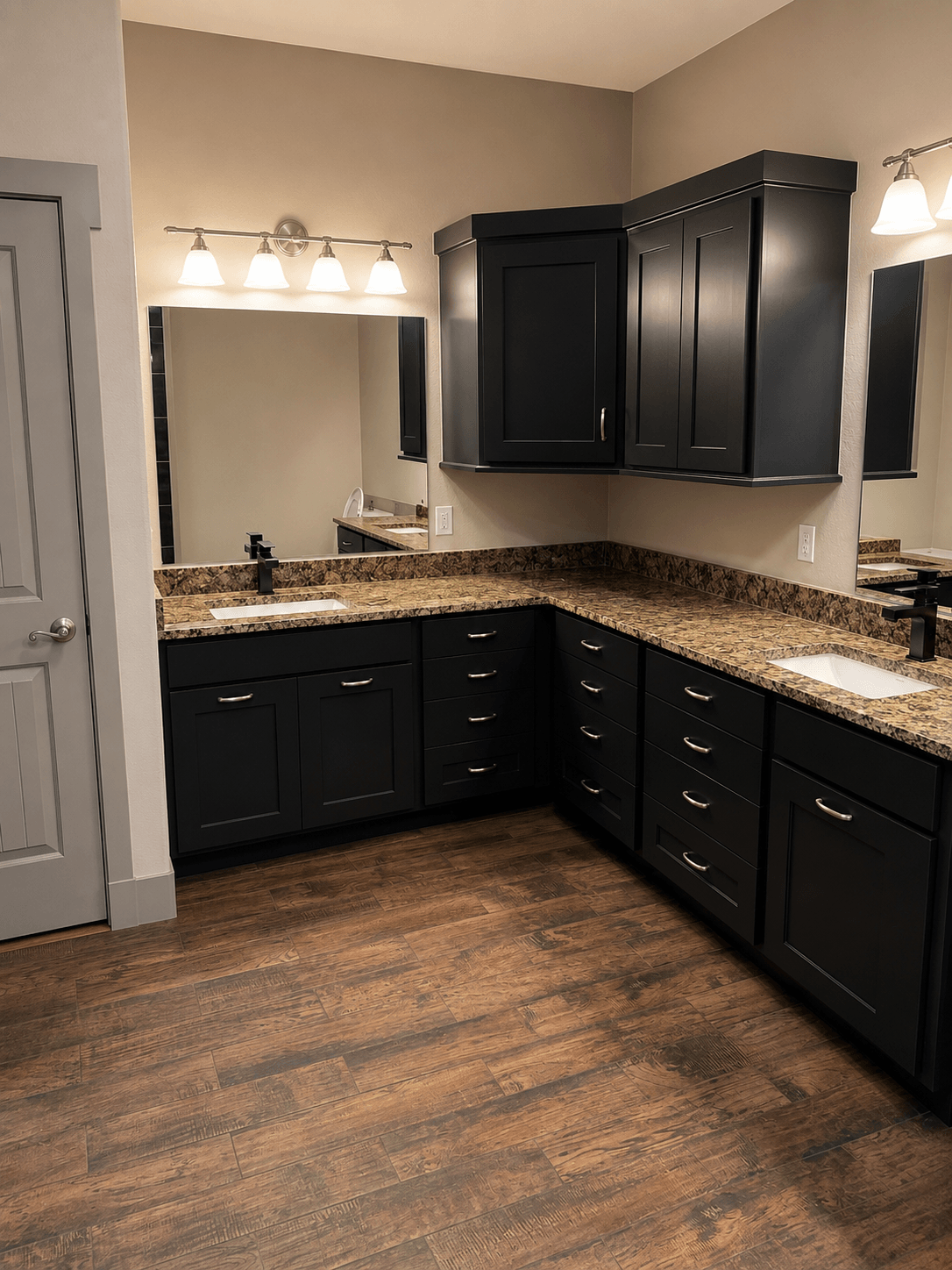 Bathroom vanity with dark cabinets, granite countertop, mirror, and lighting fixtures, wood floor, and wall-mounted cabinets.