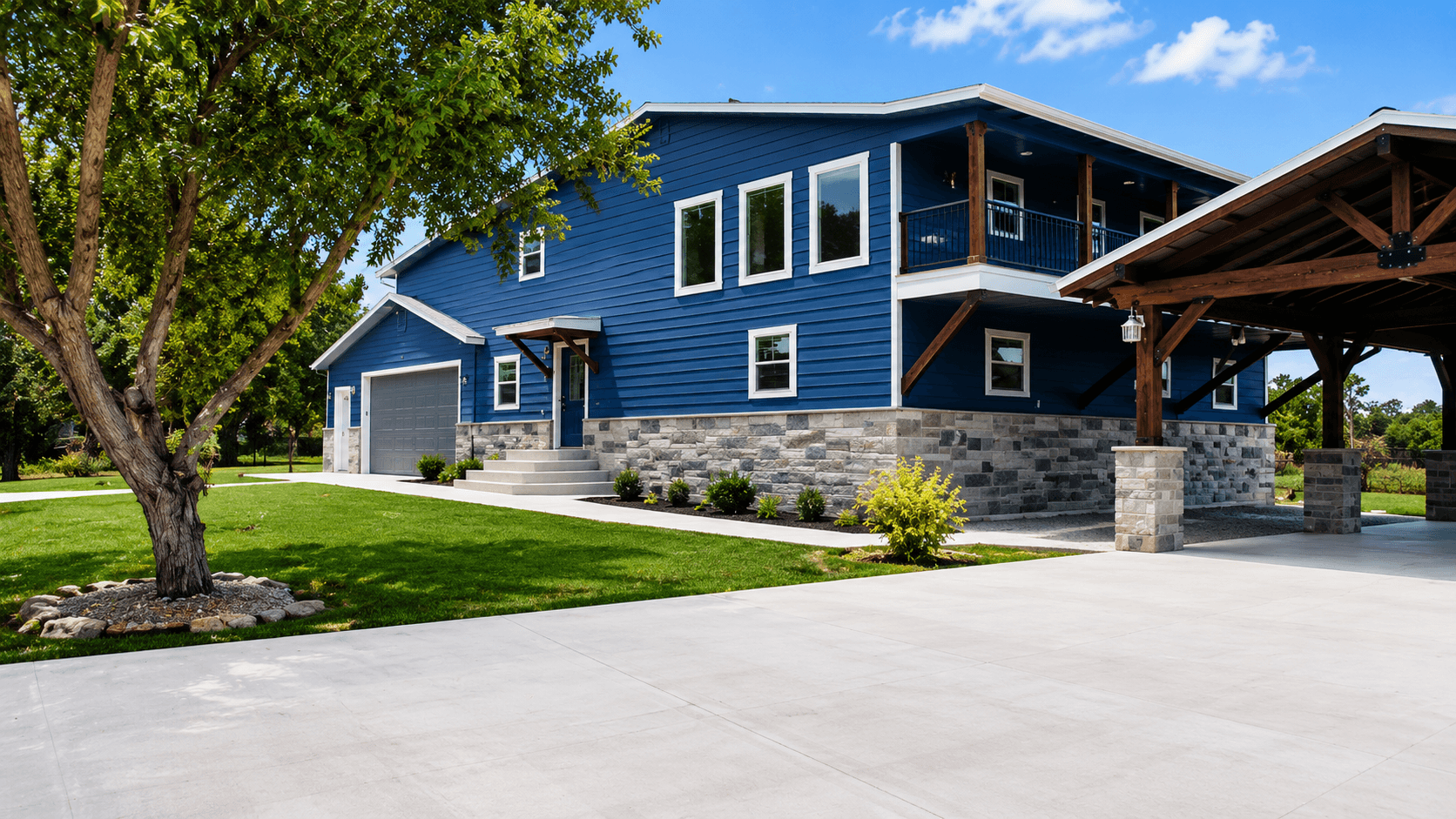Blue house with multiple windows, stone foundation, surrounded by green lawn and trees, with a driveway and a wooden carport.