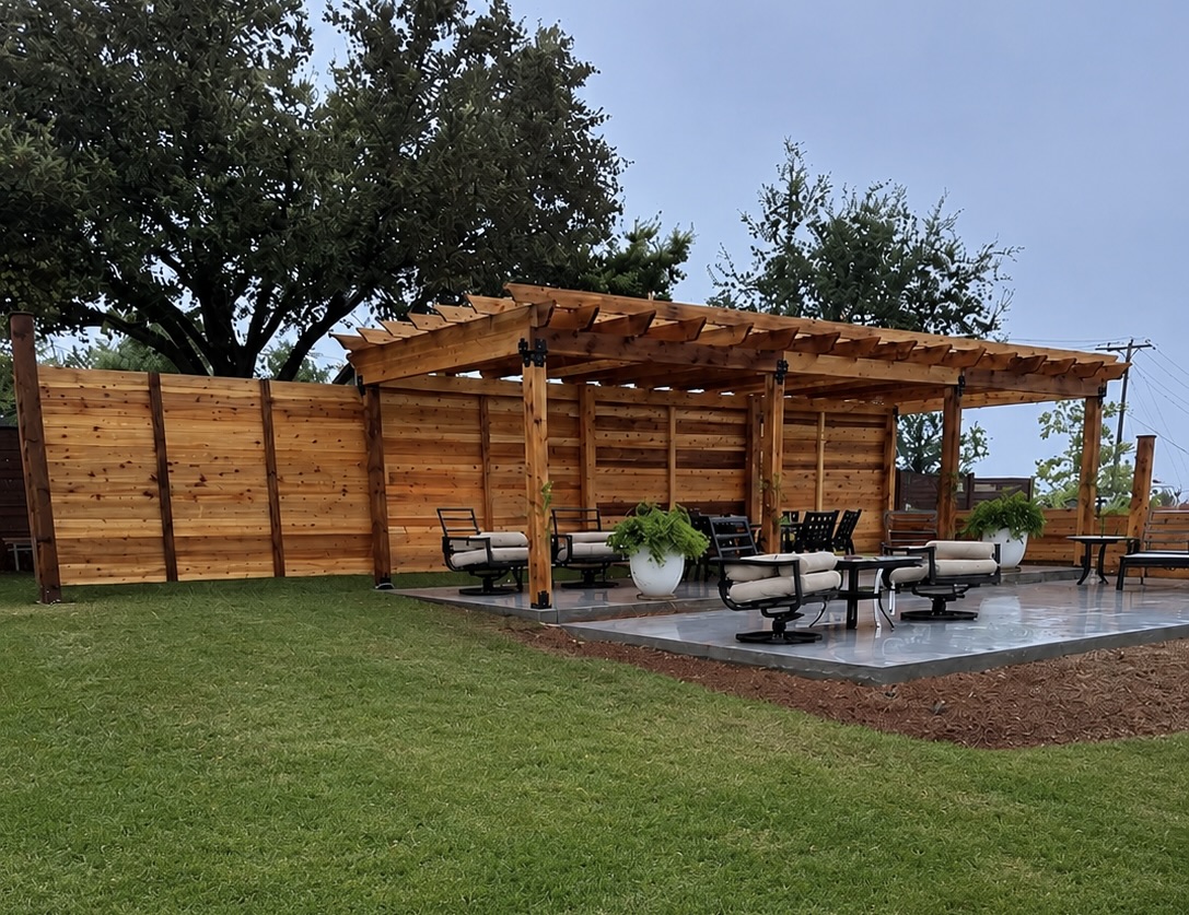 Outdoor patio with wooden pergola, seating area, and potted plants, surrounded by a wooden fence and green lawn.