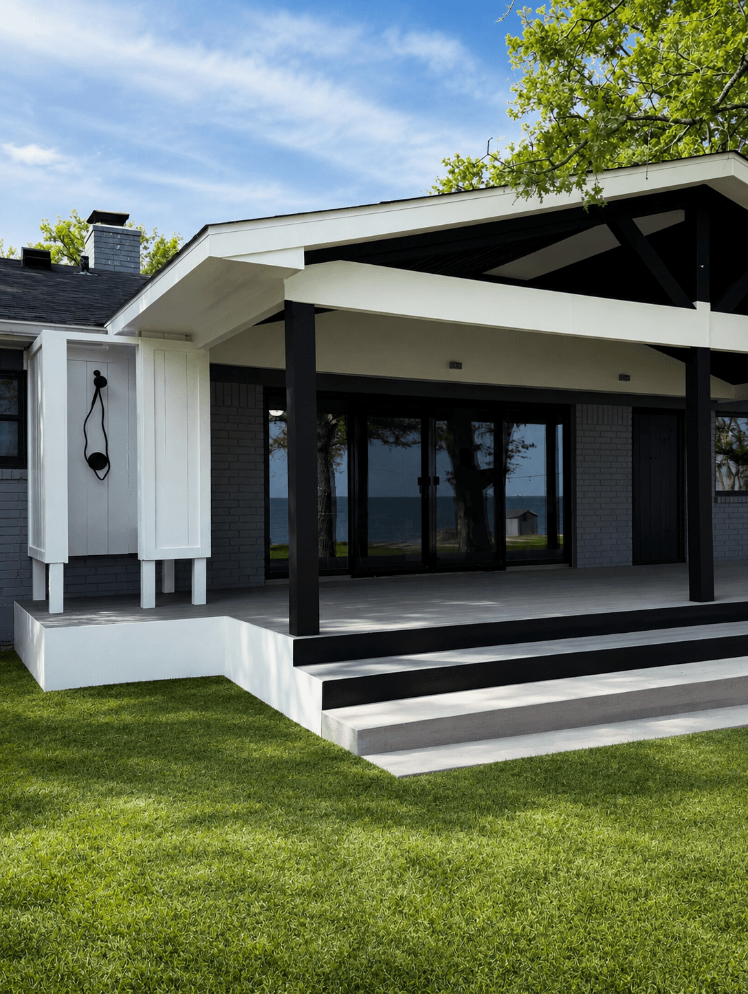 Modern house with a covered porch, black support columns, and large glass doors, surrounded by green grass and trees.