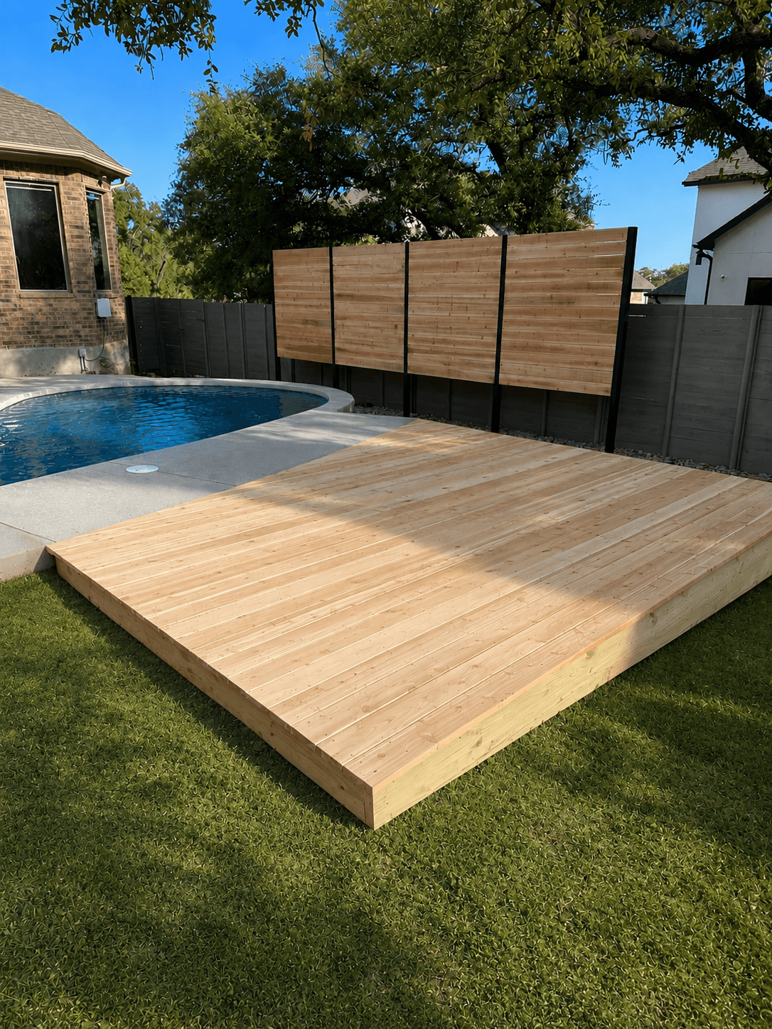 Wooden deck in backyard with privacy screens, next to a pool, surrounded by grass and trees.