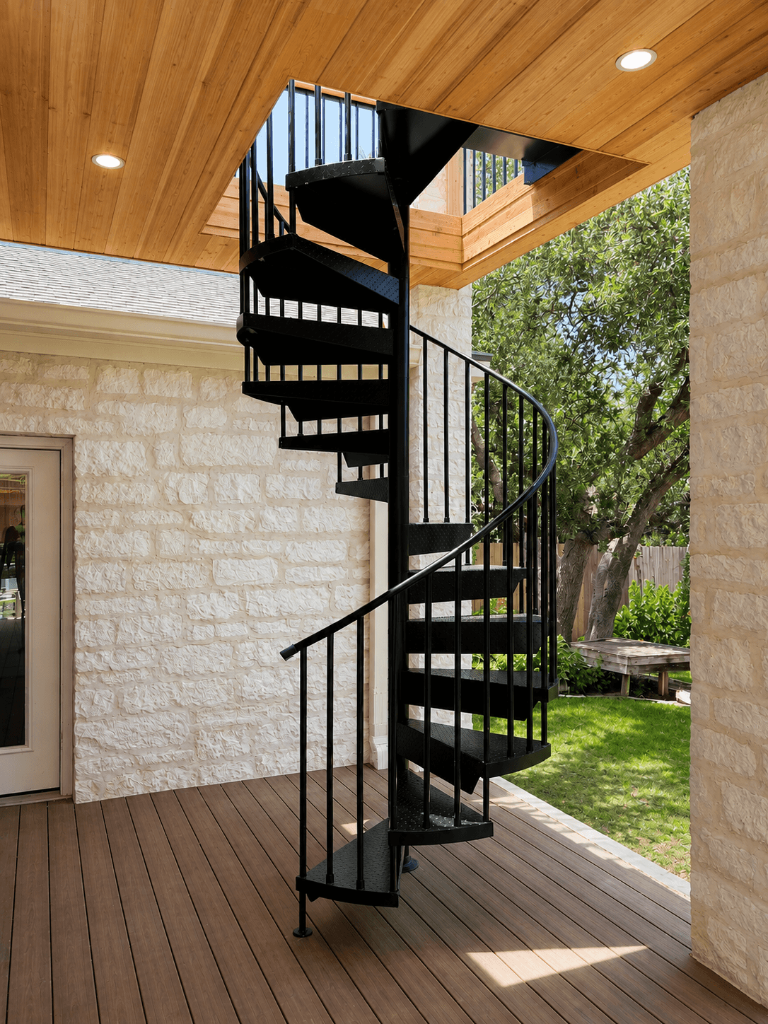 Spiral staircase with black steps and railing, leading to an upper level outside, with trees and sky visible in background.