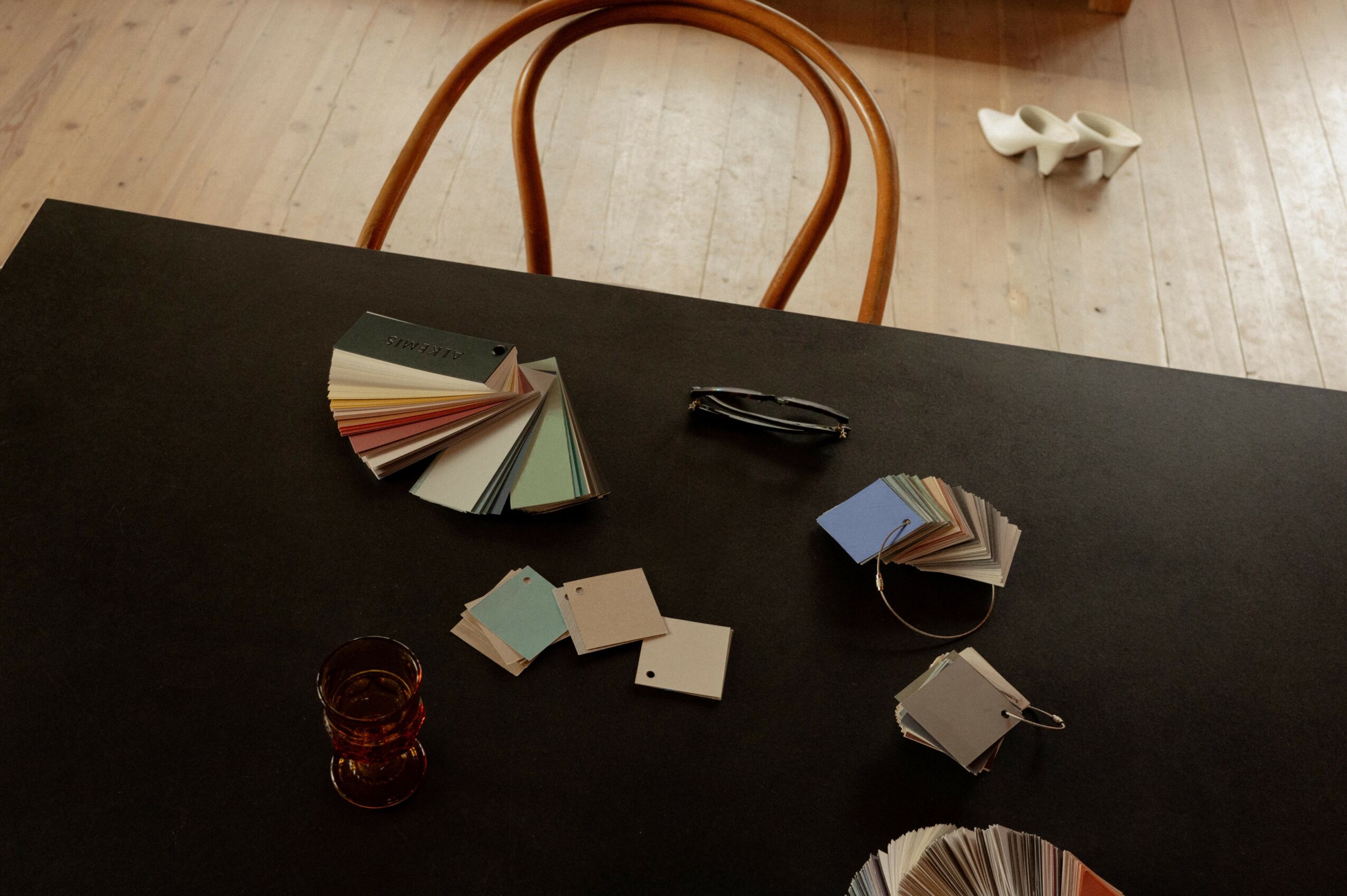 Color swatches fanned out on a black table with a small glass jar, paper clips, and a pair of glasses, viewed from above.