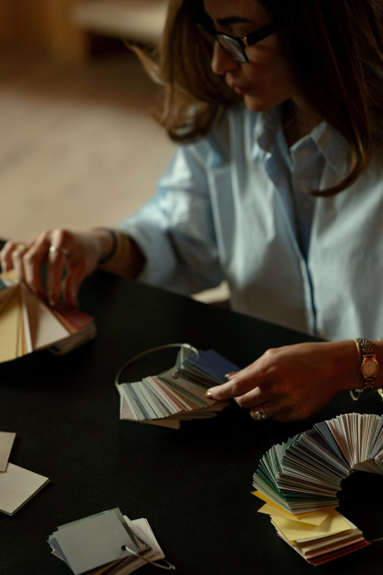 Person with glasses and long hair looking at color swatches on a table, wearing a light blue shirt.