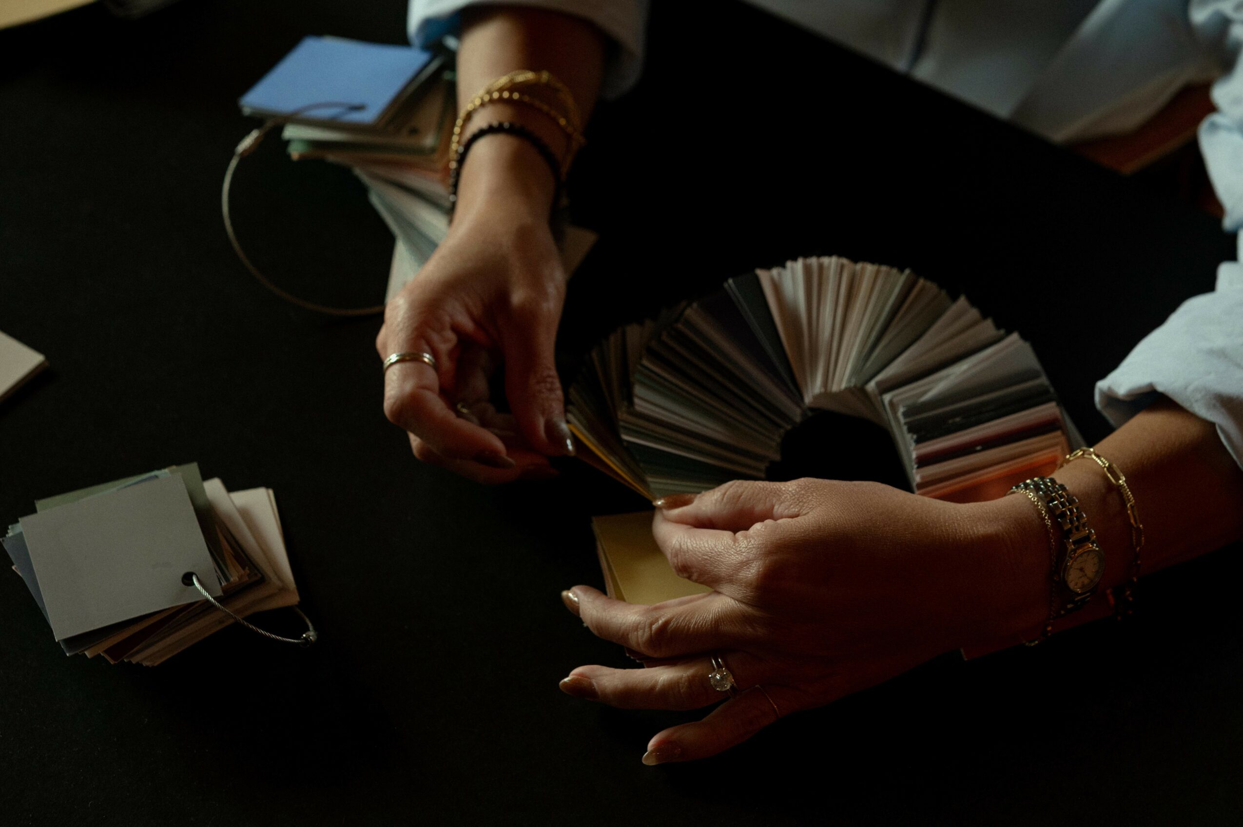 Person's hands flipping through color swatches or sample cards, with a dark background and some jewelry visible.