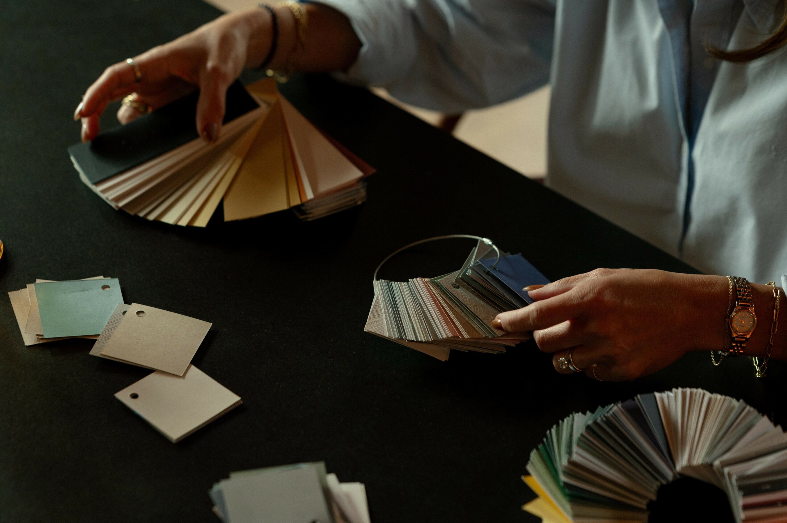 Person's hands holding color swatch fan decks, with additional swatches on dark table surface.